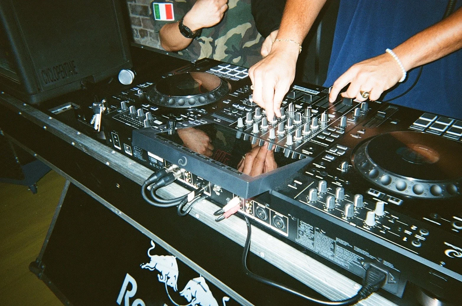 A DJ mixing music at a digital turntable with knobs and sliders, wearing a bracelet and ring, with a speaker in the background.