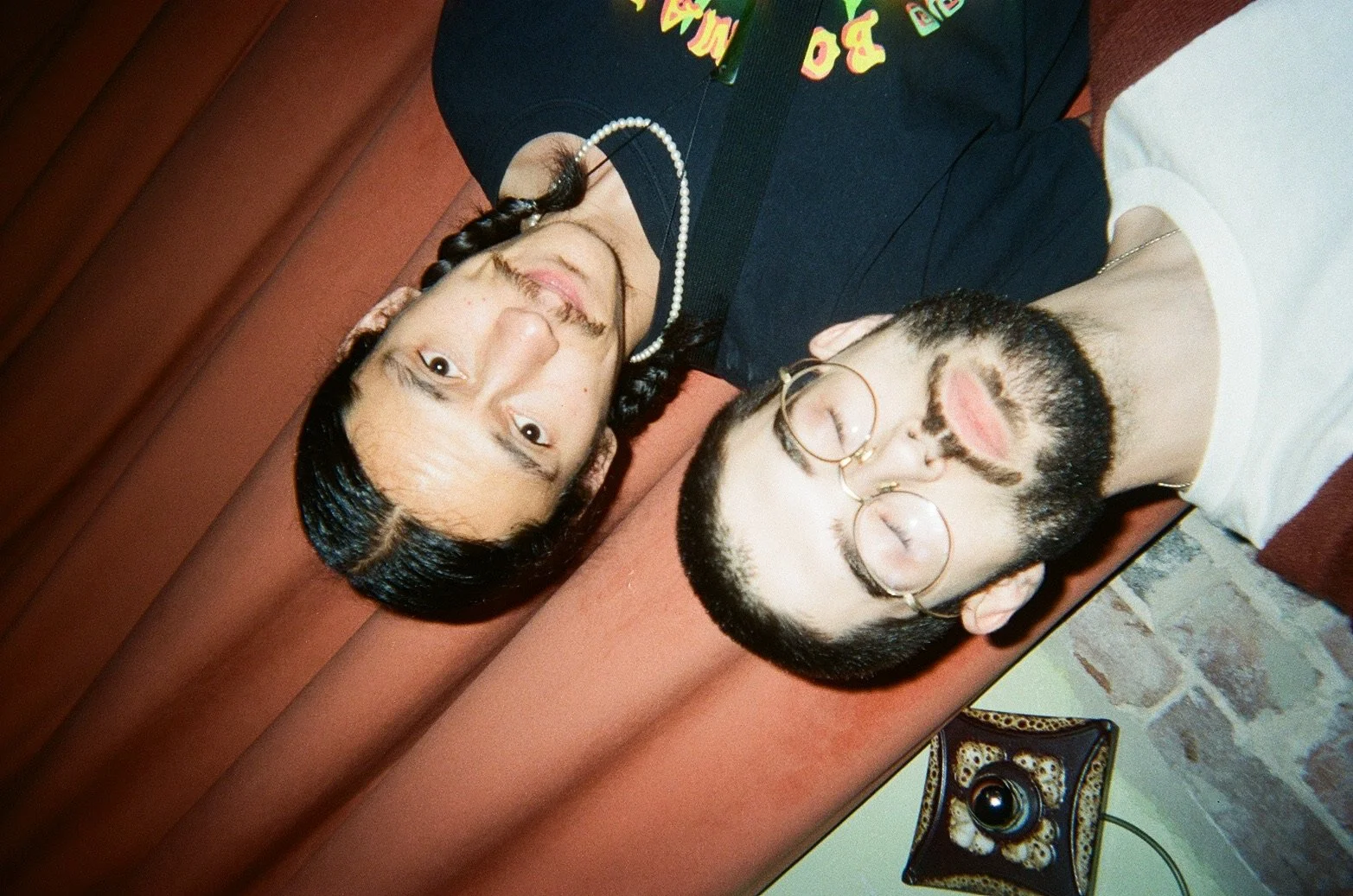 A woman and a man are lying on their backs on a brown bench, with their heads close together. The woman has long, dark hair with a braid on one side and is wearing a black top, a pearl necklace, and makeup. The man has short dark hair, a full beard, 