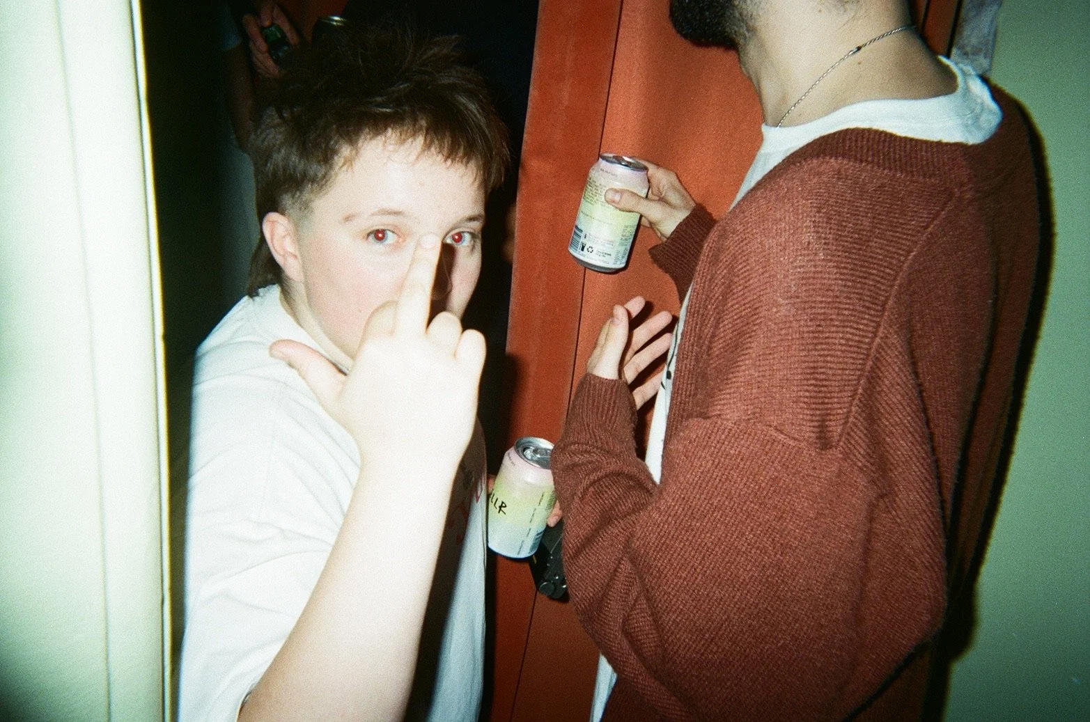 Young person with short, dark hair making a pointing gesture near their face, standing close to another person holding a soda can, both in casual clothing, in an indoor setting with a focus on their partial profiles and the soda cans.