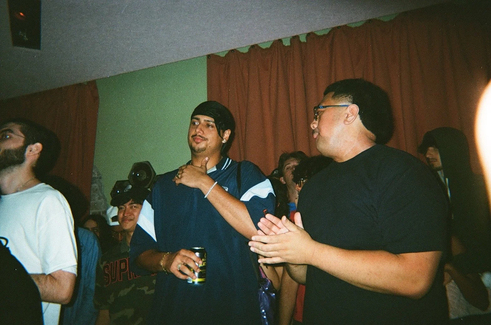 Group of people at an indoor gathering, some clapping and smiling, with a man in a dark t-shirt and glasses in focus, holding his hands together.