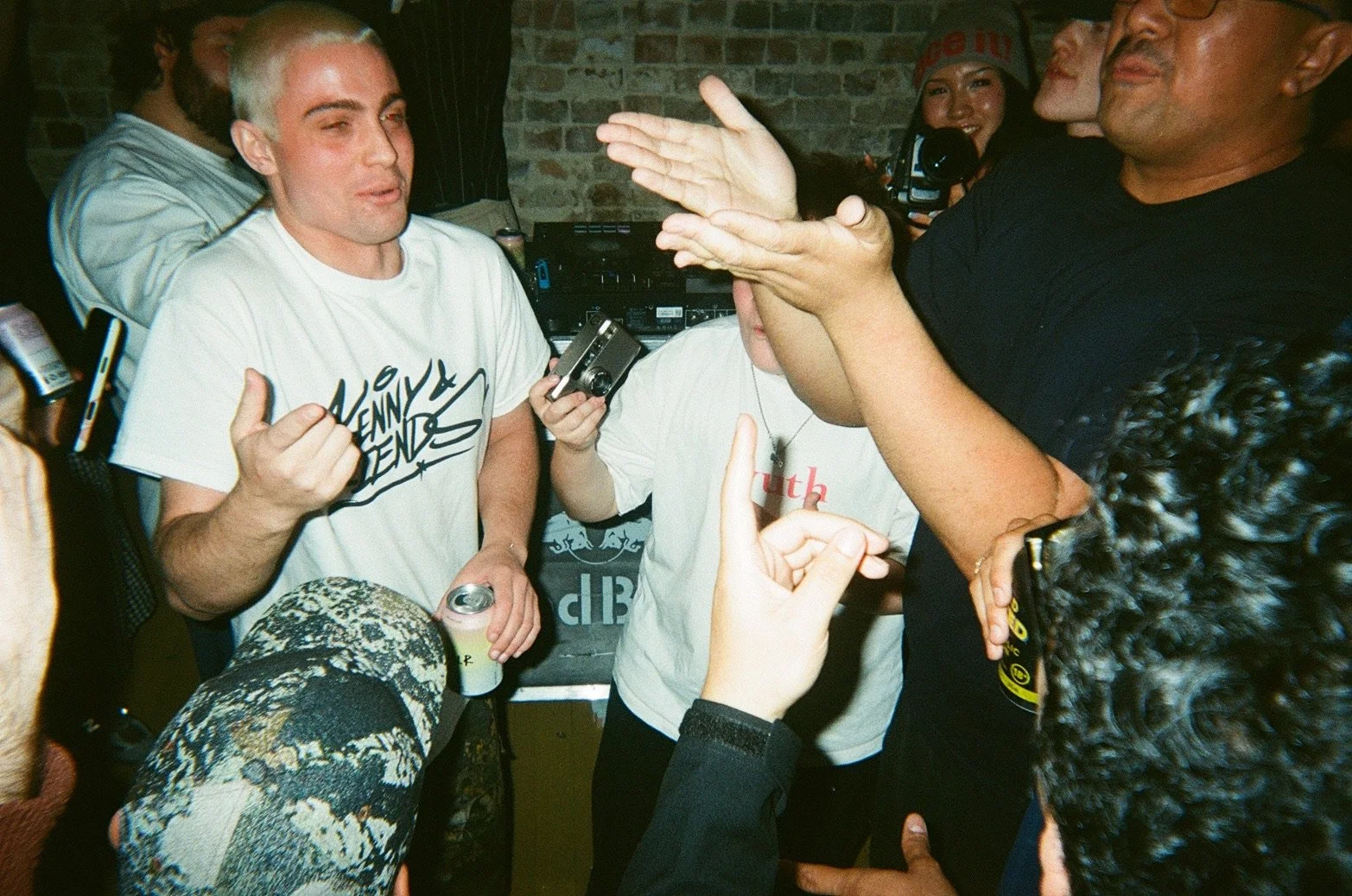 Group of people gathered in a room with brick walls, engaging in conversation and making hand gestures. One person is holding a can, and another is holding a camera.