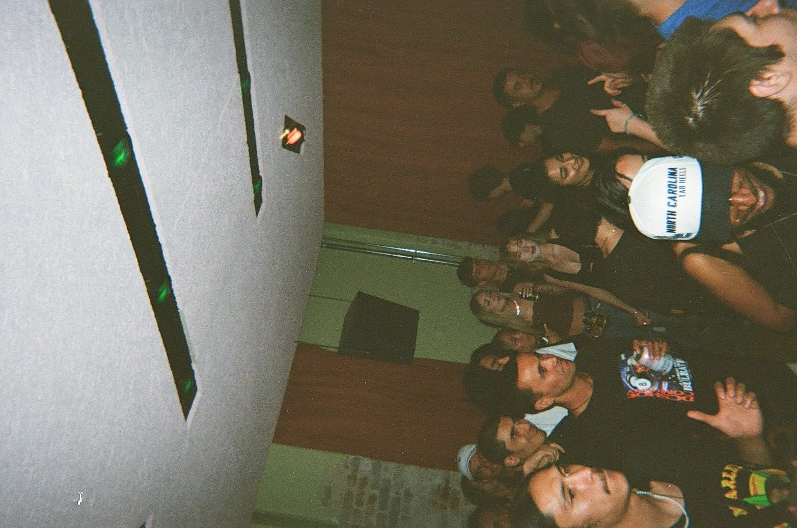 Group of young people at a party or social gathering, some holding drinks, standing in a room with wood-paneled walls and a stone wall, facing the camera, many smiling and posing.