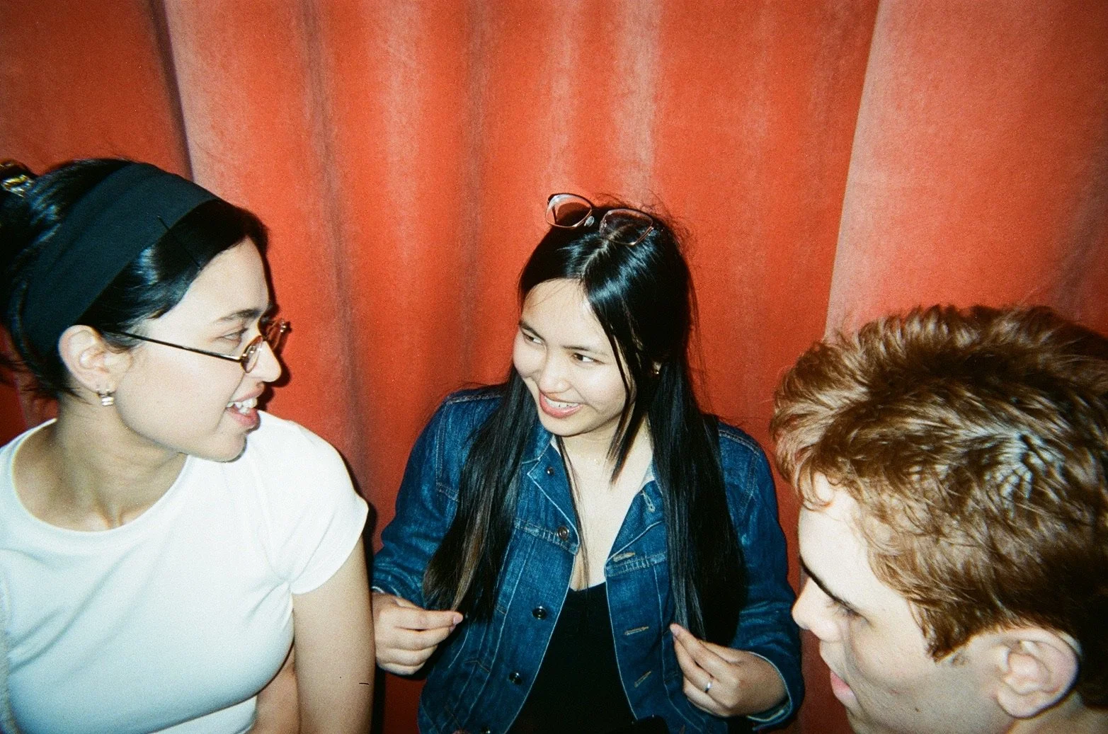 Three young adults engaged in conversation indoors, with a red curtain in the background. The woman on the left wears glasses and a white shirt, the woman in the middle wears a denim jacket and glasses on her head, and the man on the right has brown 