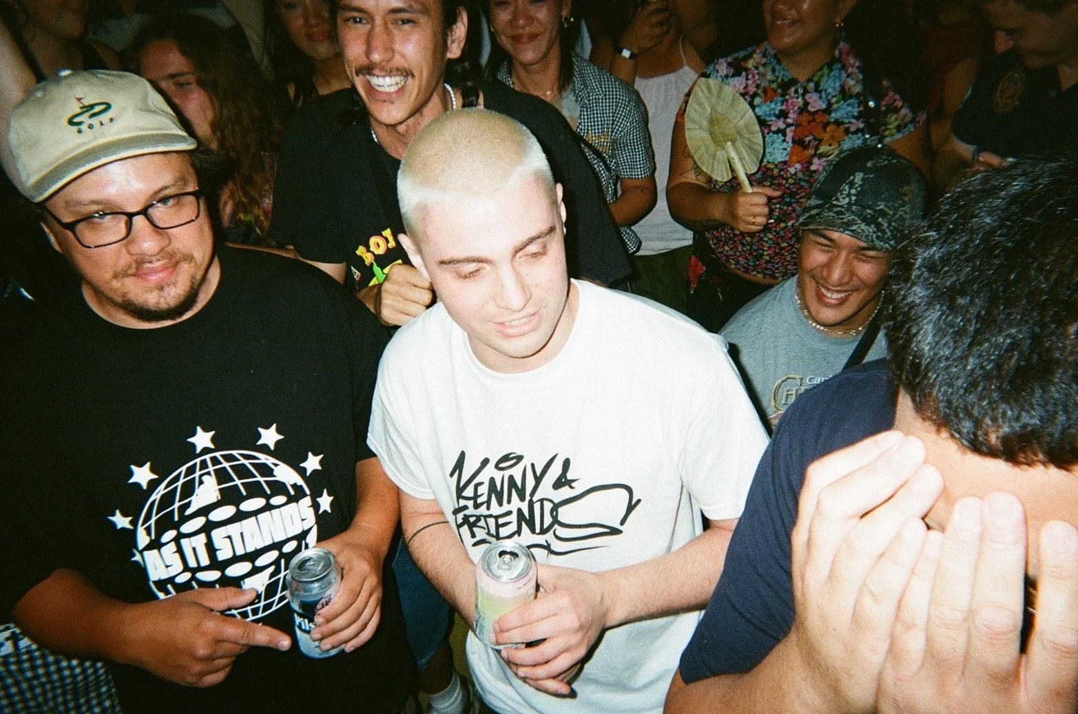 People at a crowded gathering, some smiling and holding drinks, with a man in a black shirt with a globe and stars logo and a man in a white Kenny & Friends shirt in the center, surrounded by others who are cheering and talking.