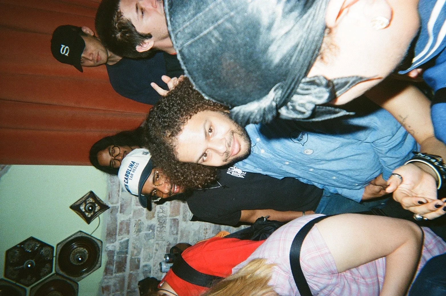 Group of people at a party or social gathering, with some making peace signs and looking at the camera, against a background featuring brick wall and speakers.
