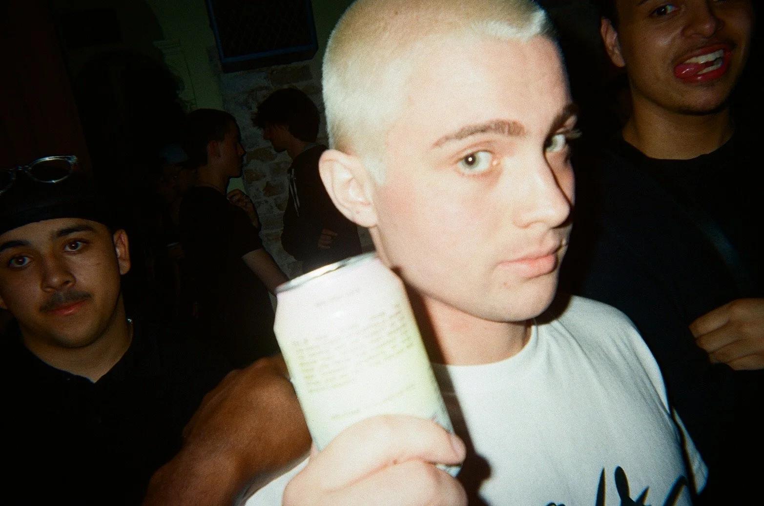 Young person with short blonde hair holding a can, surrounded by other people at a social gathering or party.