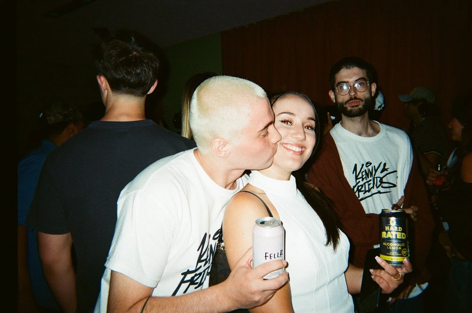 Group of young adults at a party, some holding canned drinks, with a woman smiling in the center, a man kissing her cheek, and others in the background.