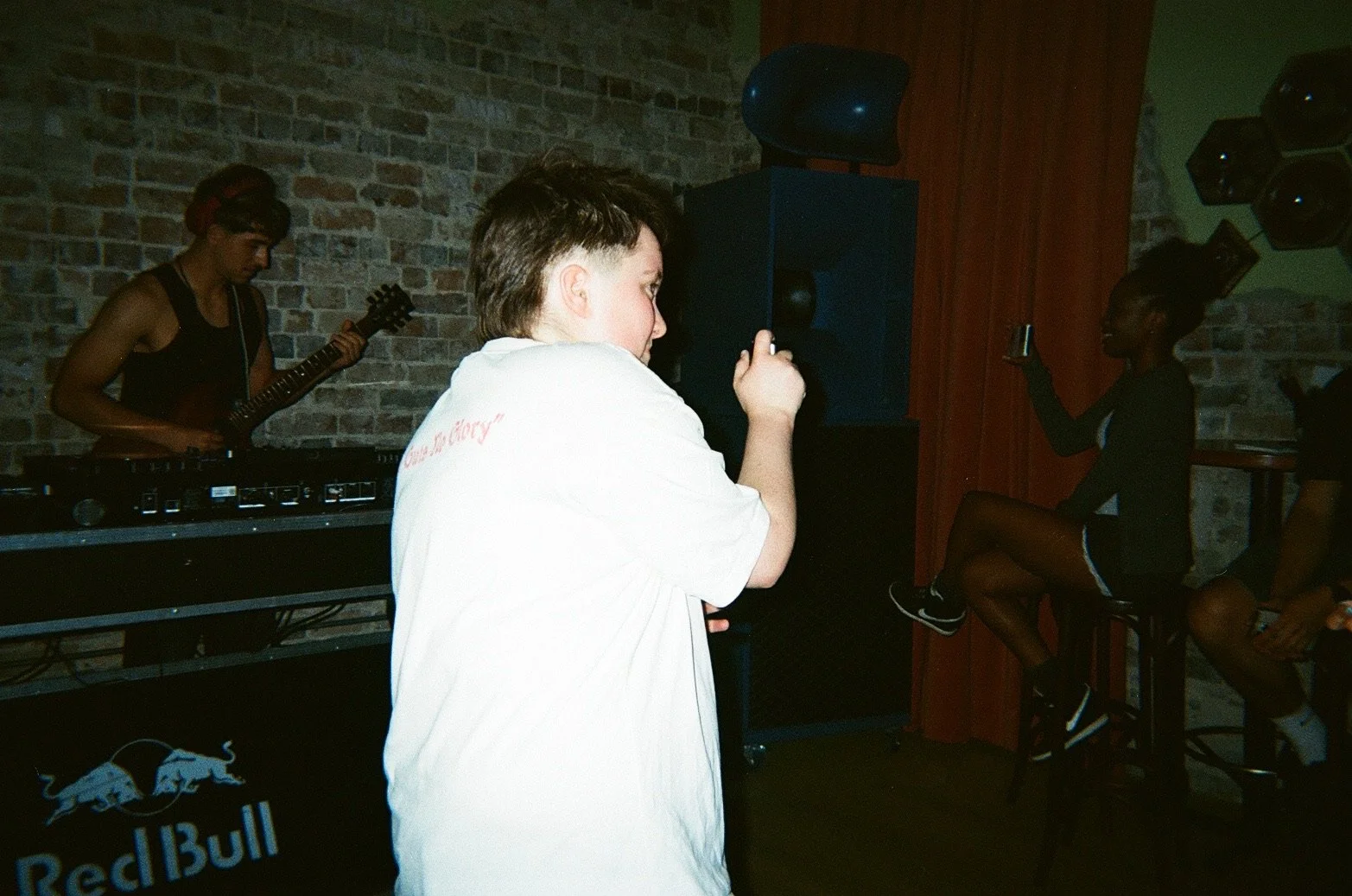 A young person with short, dark hair dancing and singing in a dimly lit room with brick walls, while a guitarist plays in the background. An audience member is sitting on a stool, holding a drink and watching the performance.