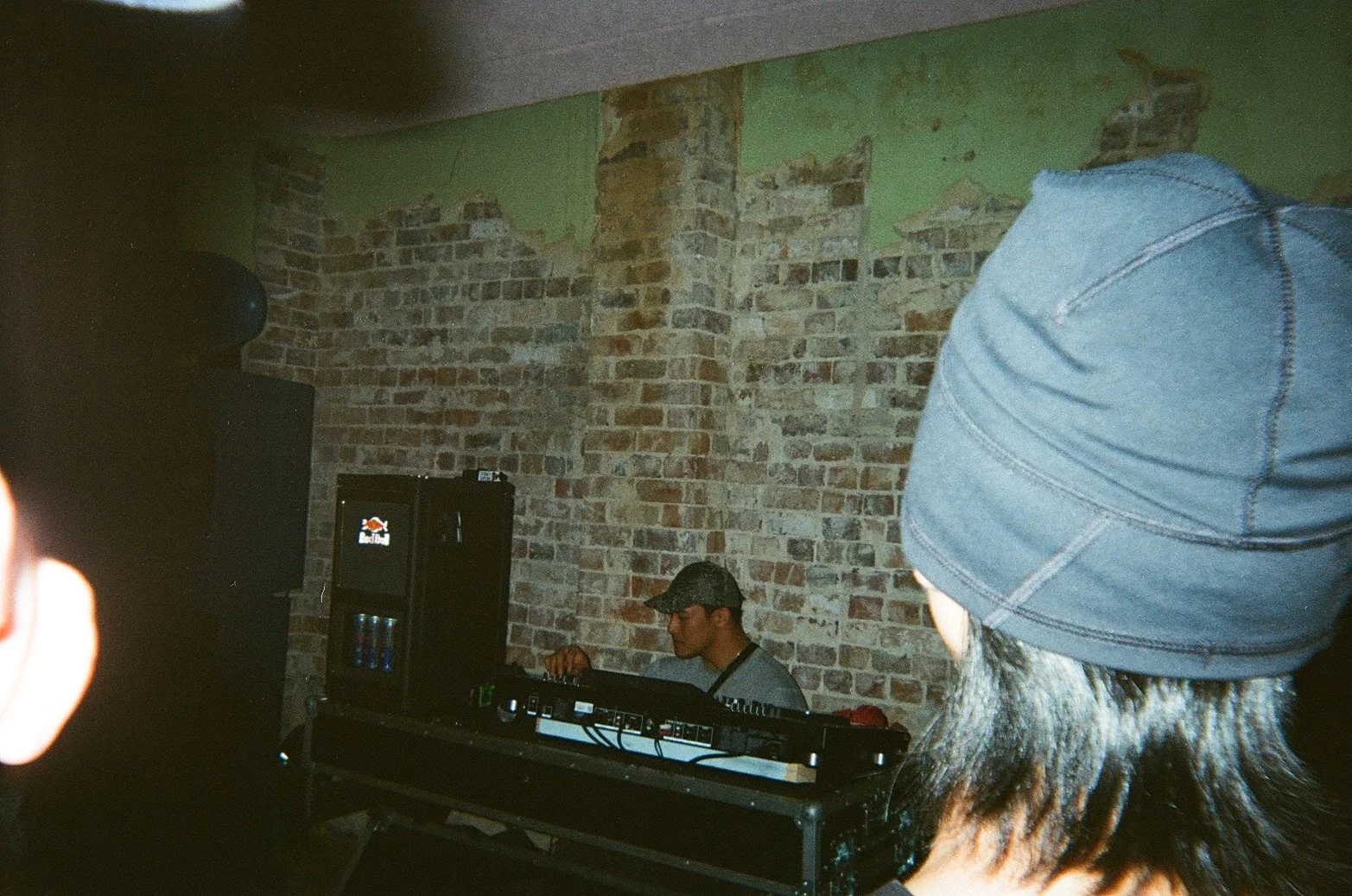 A DJ wearing glasses and a camouflage cap performs on a turntable setup in front of a exposed brick wall, with people watching, in a dimly lit indoor venue.
