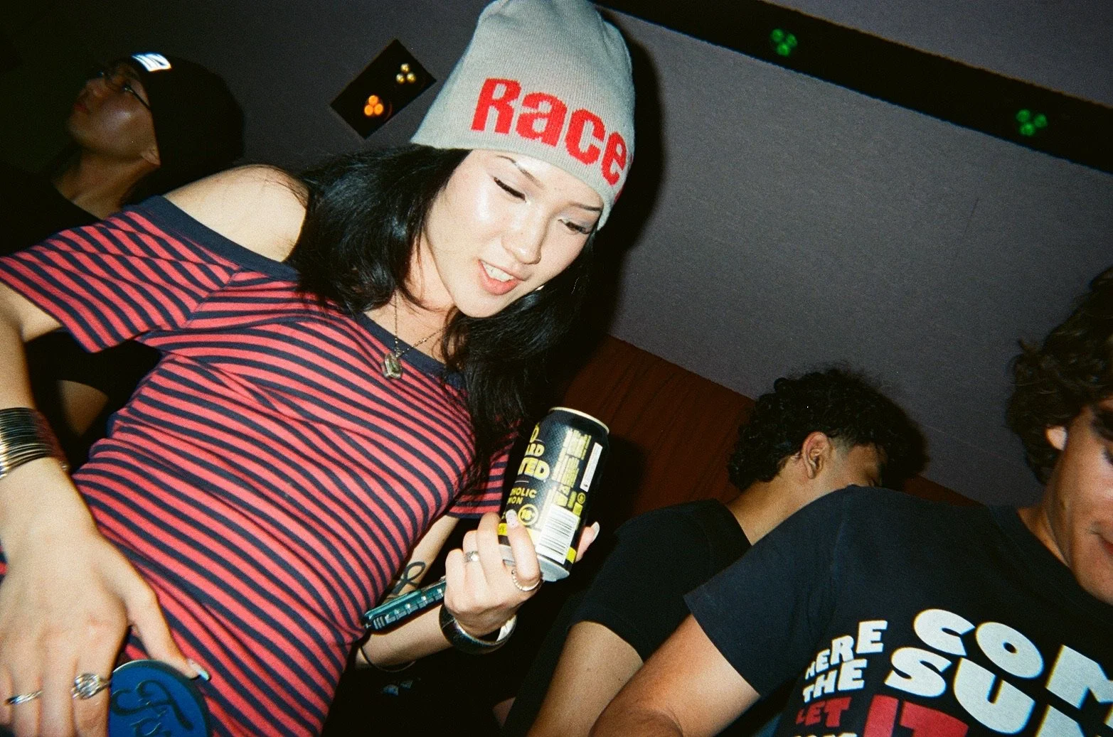 A young woman with dark hair wearing a gray beanie with red text, a red and blue striped shirt with shoulder cutouts, and various jewelry, holding a can and looking down at her phone, in a dimly lit social setting with other people around.