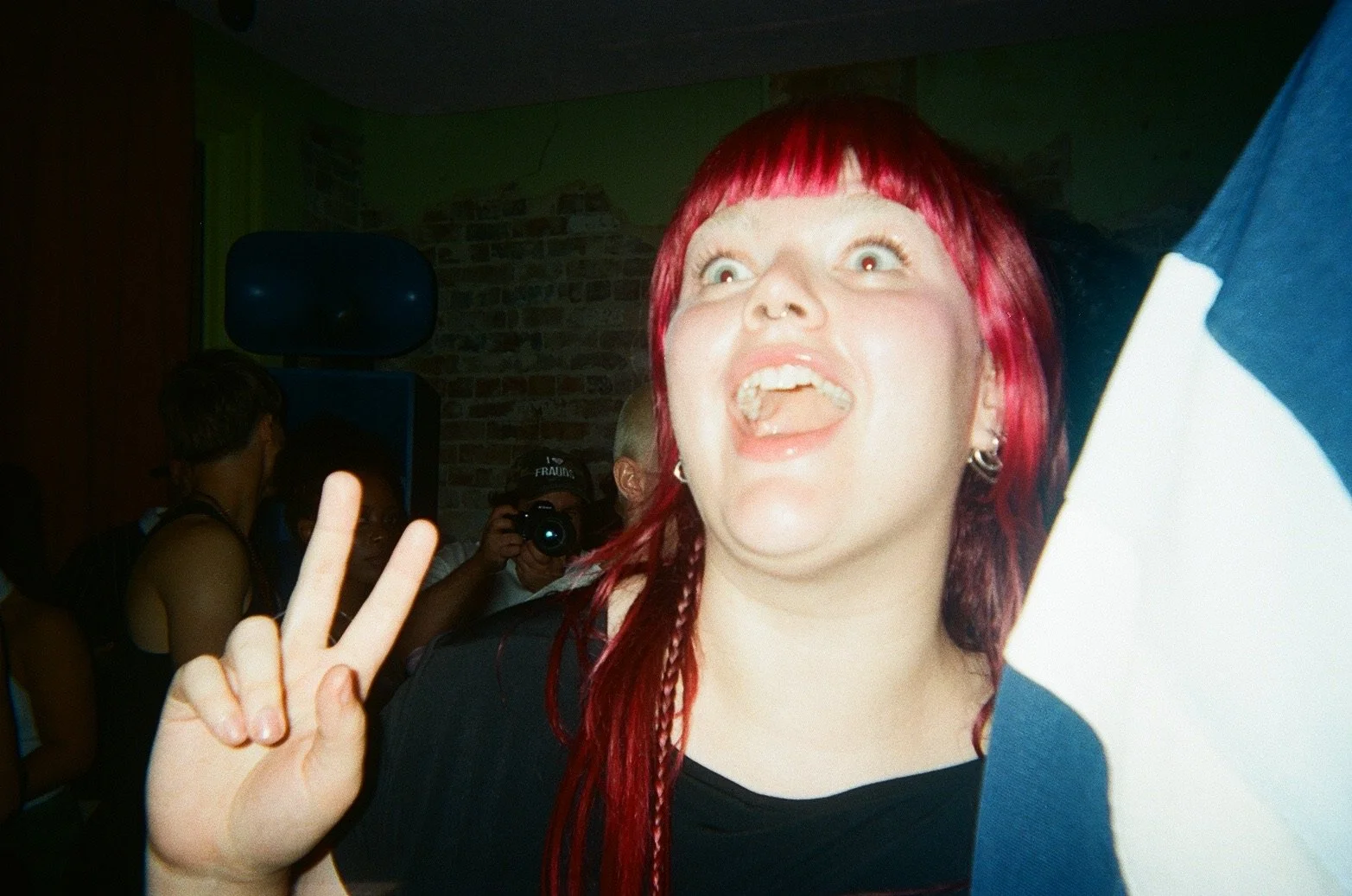Close-up of a woman with red hair making a peace sign and sticking her tongue out at a party or event with other people and a man taking a photo in the background.
