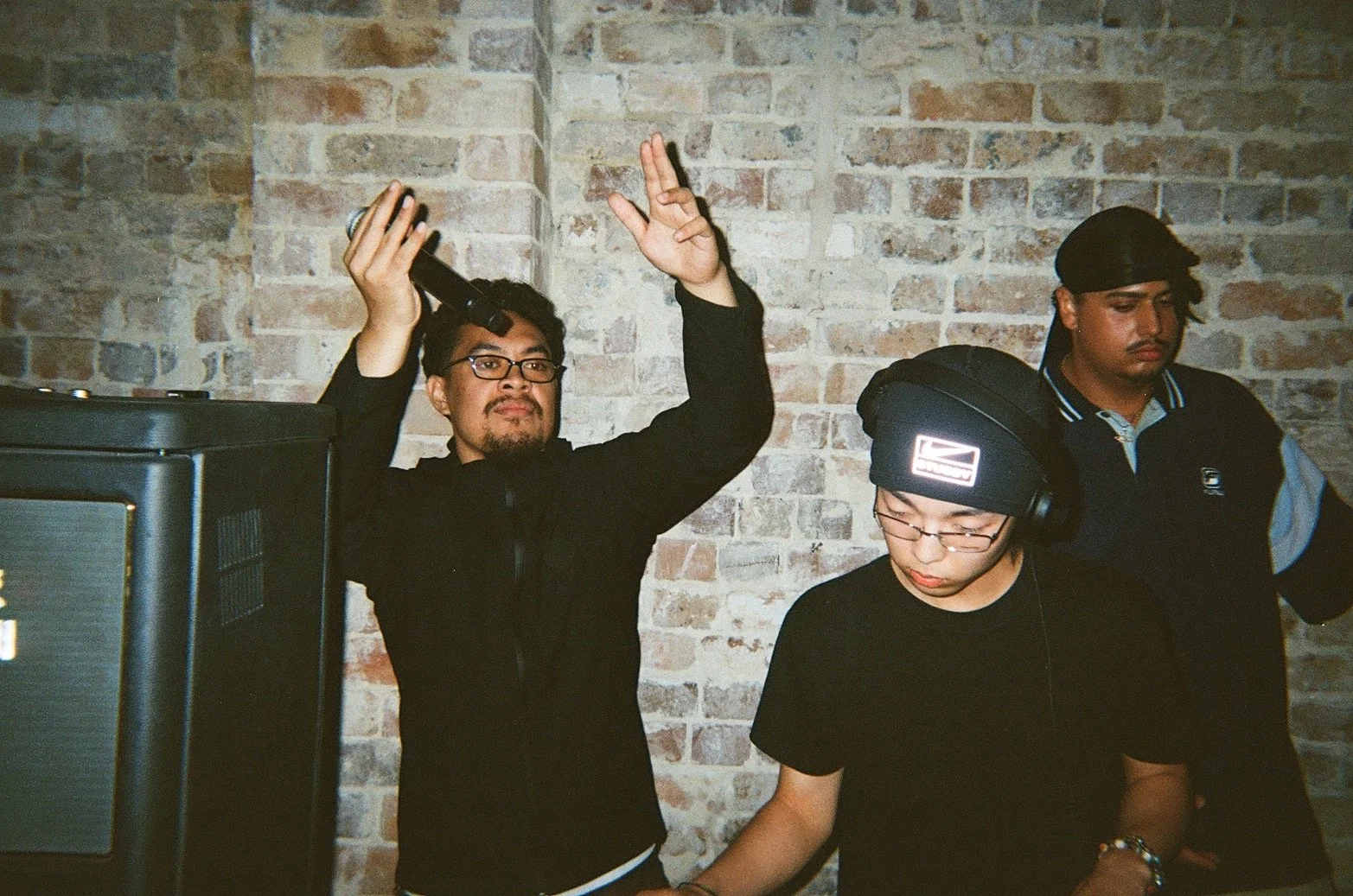 Three young men are standing in front of a brick wall. The man on the left is holding a microphone and raising his right hand with a peace sign; he is wearing glasses and a black jacket. The man in the center is looking down at a device, wearing glas