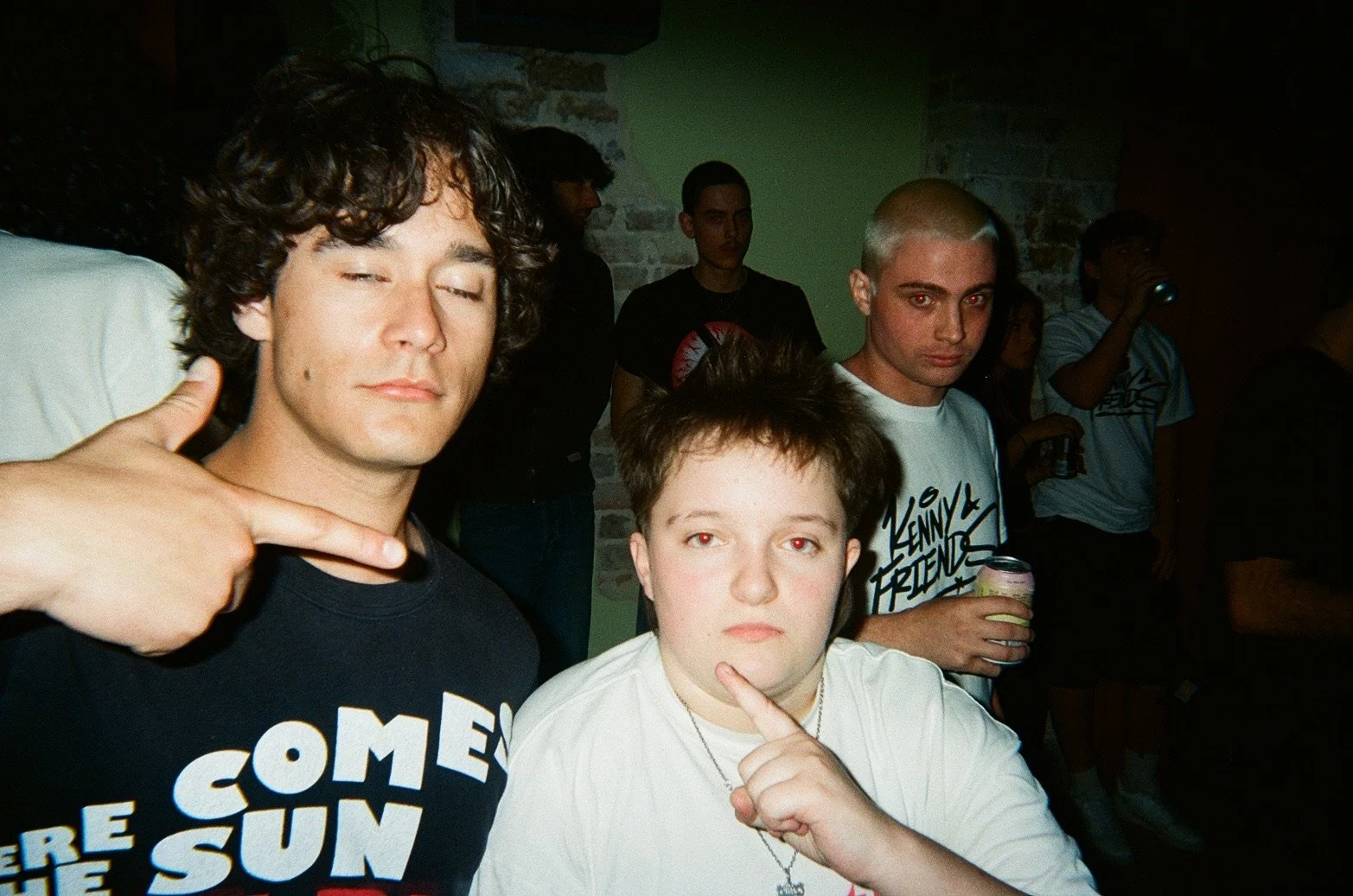 Three young people posing together at a party or gathering, with a dimly lit background and guys holding drinks, all making stylized gestures with their hands.