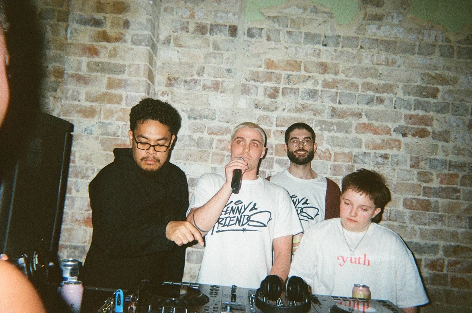 Four people stand behind DJ equipment against a brick wall. The person on the left has curly black hair and glasses, wearing a black shirt. The second person from the left has short platinum blonde hair, speaking into a microphone, wearing a white t-