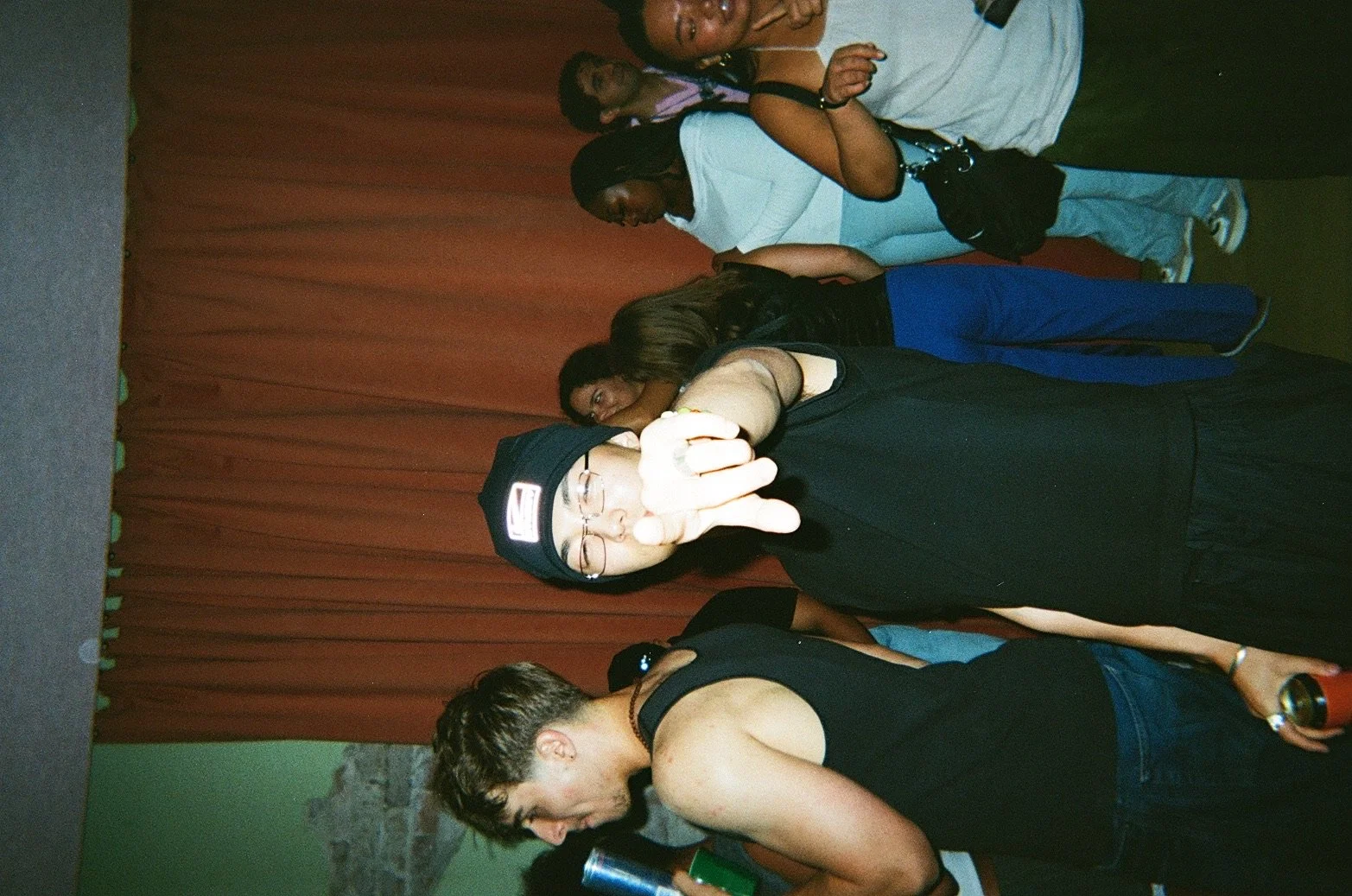 A group of people at a social event with a brown curtain background, including a person in a black hoodie giving a peace sign and others standing around.