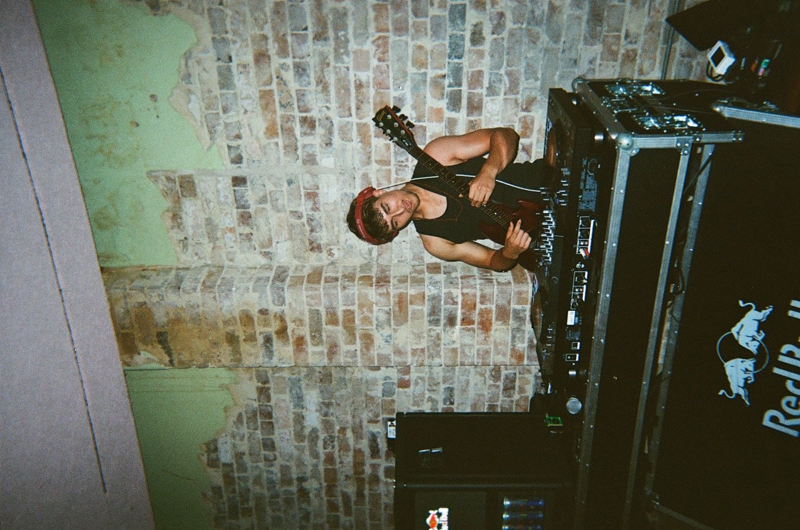 A woman with short hair, wearing a black sleeveless top and a red headband, is playing an acoustic guitar while standing behind DJ equipment on a table. The background features an exposed brick wall with patches of peeling green paint.