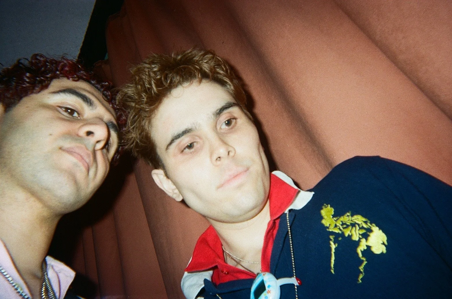 Two young men taking a selfie together, one with curly dark hair and the other with light brown, wavy hair. They are indoors against a reddish-brown background.