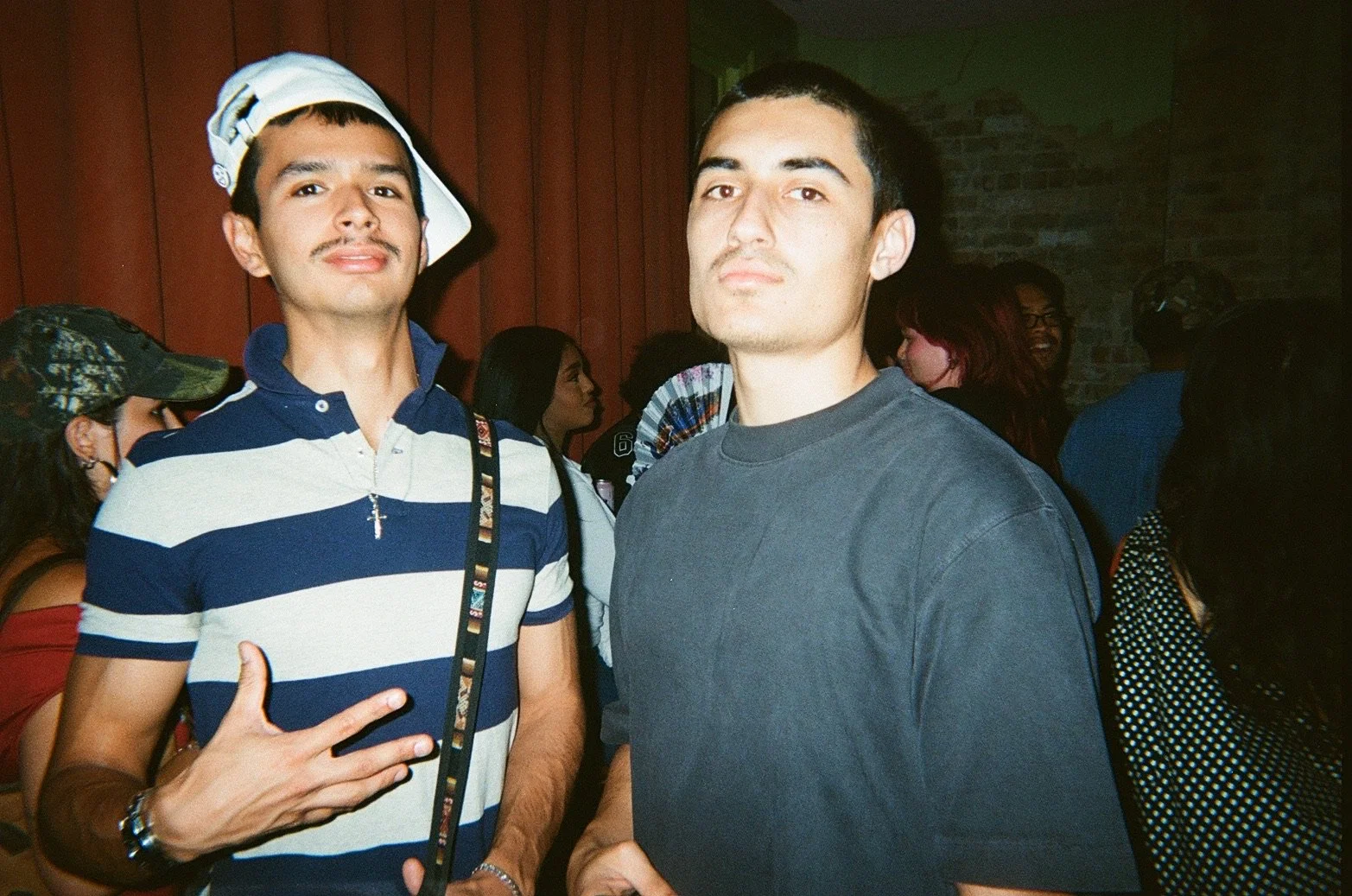 Two young men posing at a social event; one is wearing a striped polo shirt with a cap on his head and a cross necklace, making a hand gesture; the other is in a plain dark T-shirt, standing with his hands clasped in front of him. Several people are 