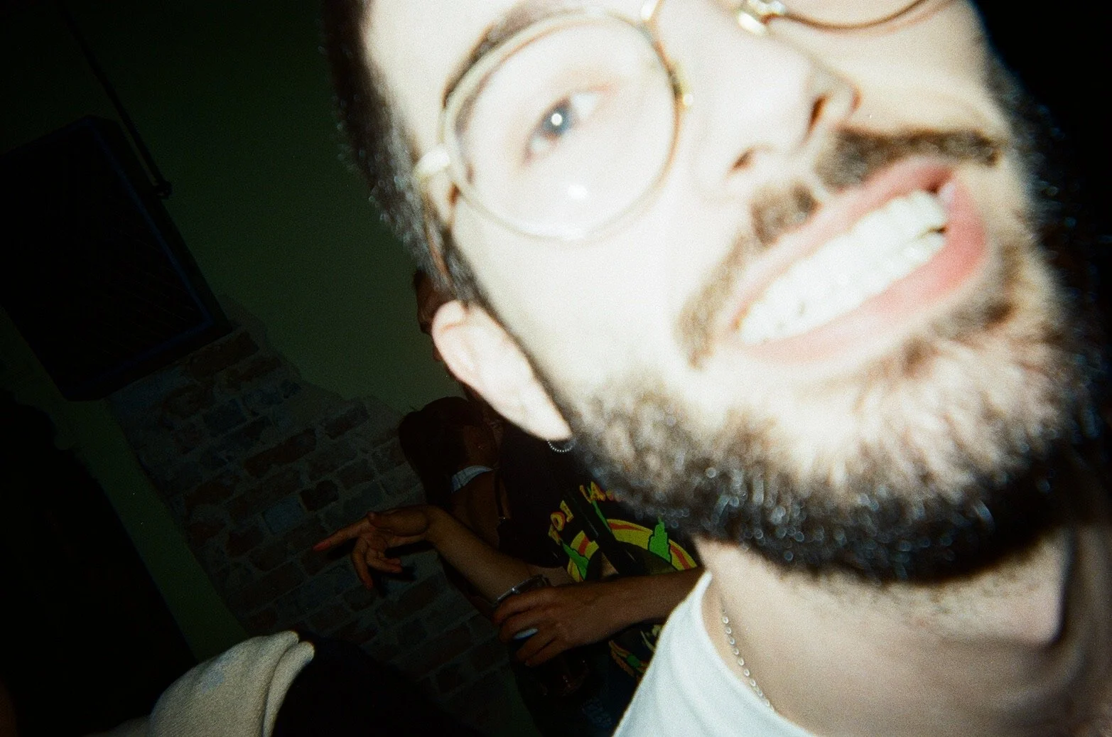 Close-up of a smiling man with glasses and a beard, showing his teeth, while someone in the background is holding a drink and making a gesture with their hand inside a dimly lit room.