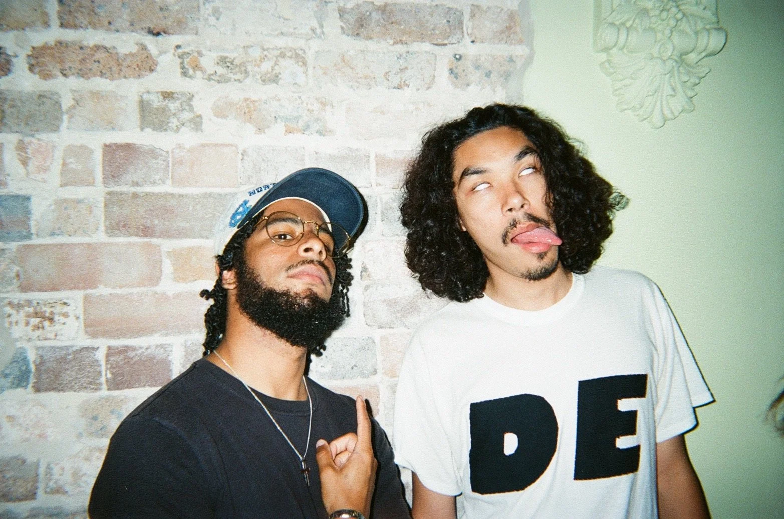 Two men standing in front of a brick wall; one with glasses and a cap, making a hand gesture, and the other with curly hair and a white t-shirt with the letters 'DE', making a face with tongue out.
