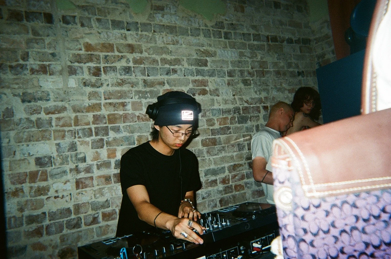 A person DJing at a club with a brick wall background, wearing glasses, a black beanie, and equipment in front of them. Two other people are visible in the background, standing and looking down.