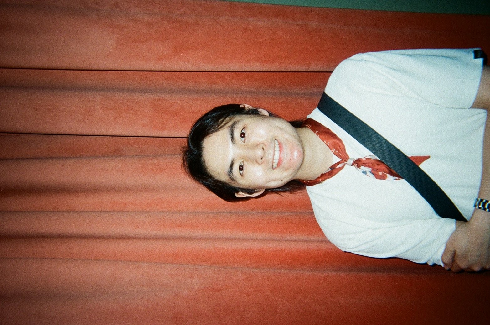 A young man with dark hair, wearing a white shirt with a black shoulder strap and a red neckerchief, smiling and looking up, standing in front of a vertical, reddish-brown wooden background.