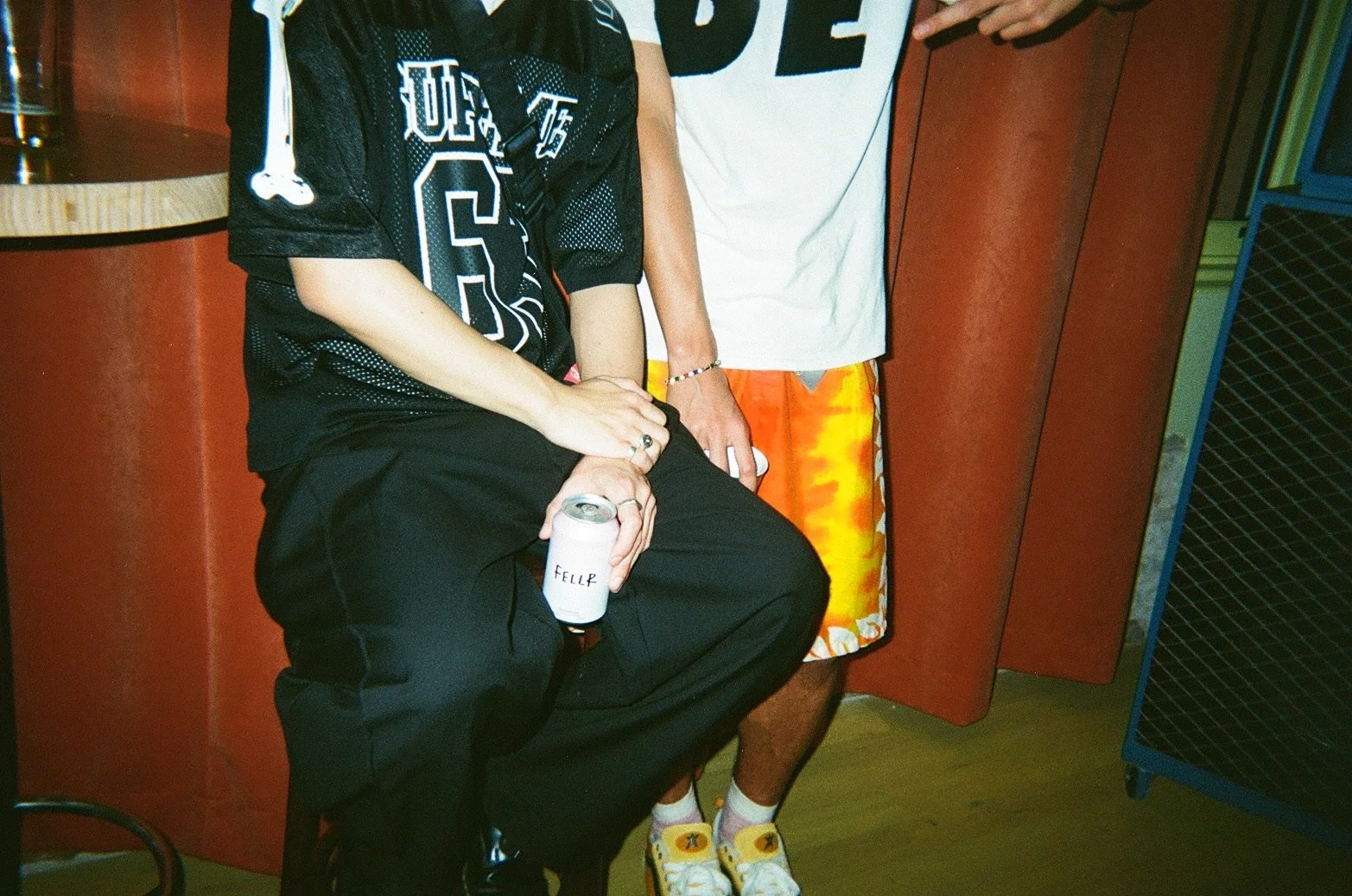 Close-up of two young men standing next to each other, one sitting on a bench holding a drink can labeled 'FELLIP'. The person on the left is wearing a black sports jersey with white lettering and black pants. The person on the right is wearing a whi