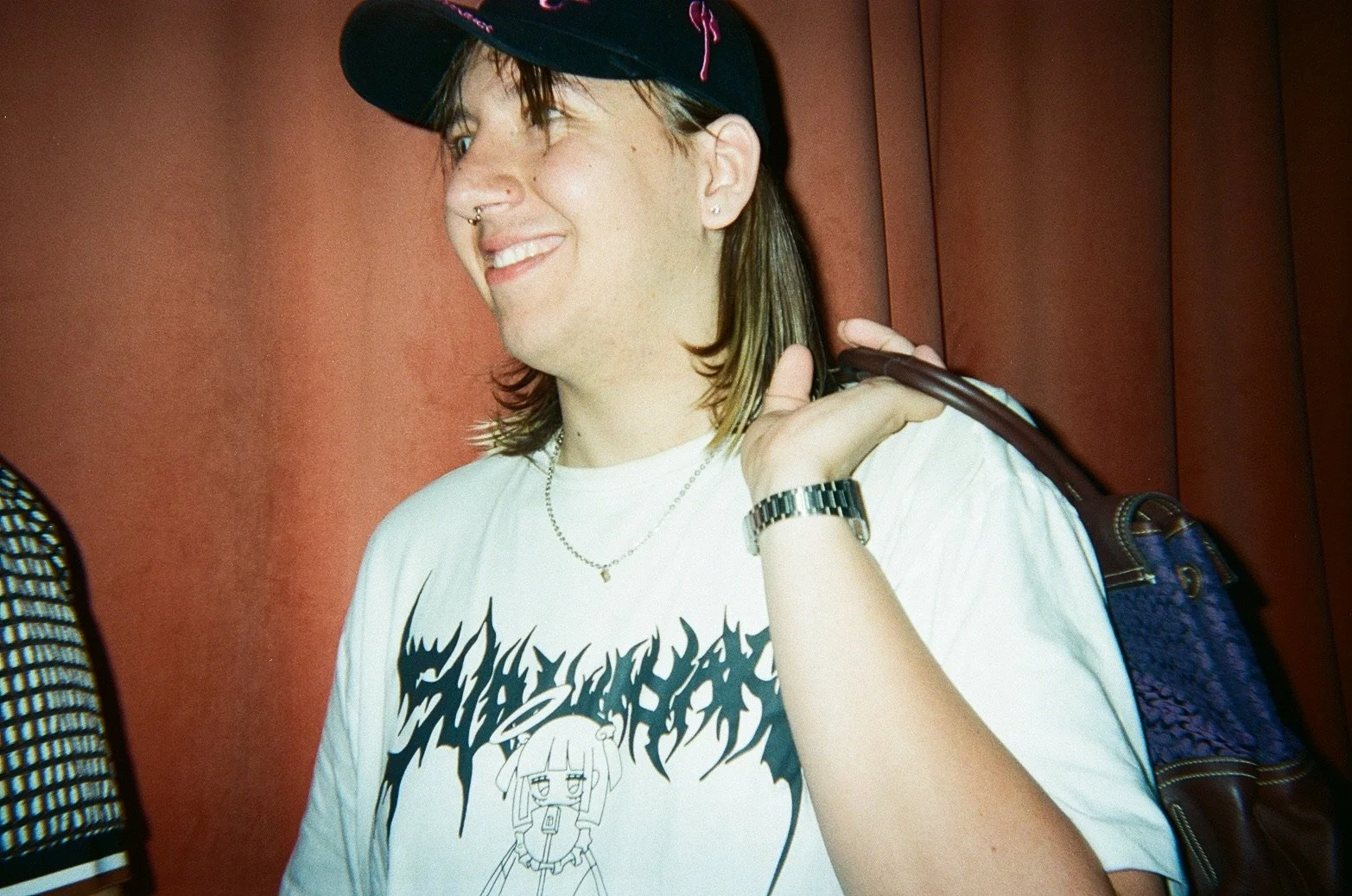 A young person with shoulder-length hair, light skin, and multiple piercings, wearing a white graphic T-shirt, a wristwatch, a necklace, and a baseball cap, smiling and carrying a bag over their shoulder against a red background.