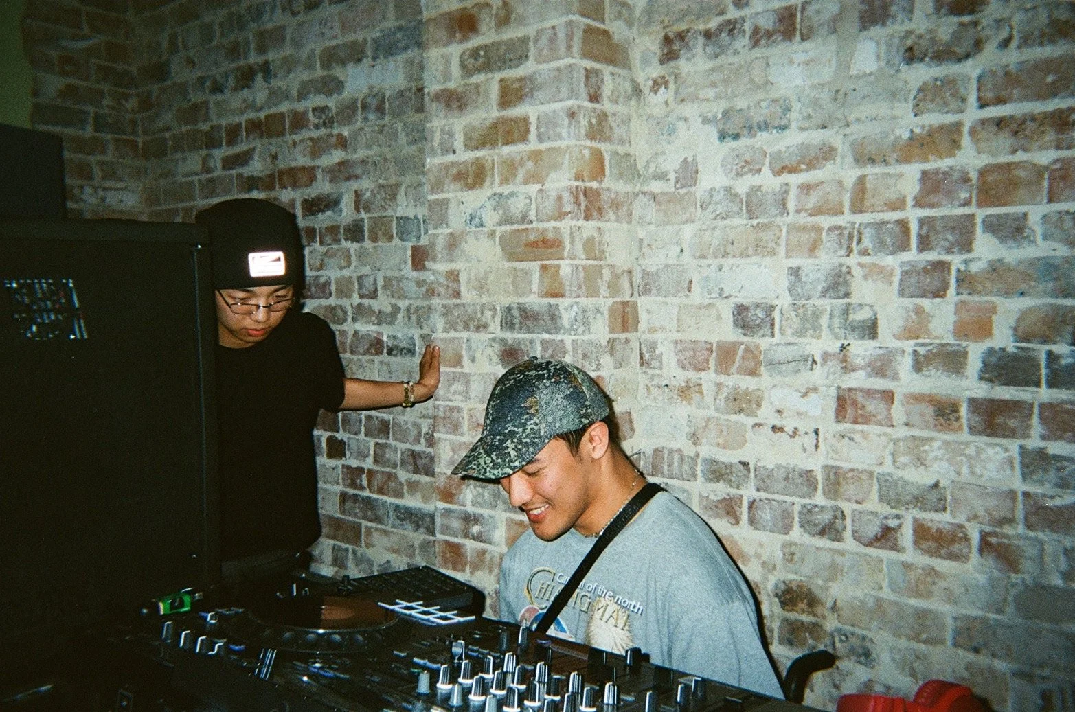 A man wearing a camo cap and a gray t-shirt is smiling, sitting in front of DJ equipment with knobs and buttons, with a brick wall in the background. A woman in black, wearing glasses and a black beanie, is standing behind him, touching the wall.