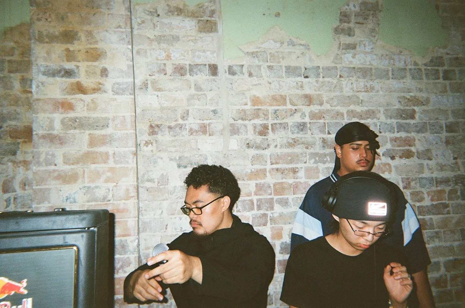 Three young people are standing against a brick wall. The person on the left is holding a microphone, wearing glasses, and has curly hair. The person in the middle, wearing a black beanie and glasses, looks down. The person on the right, with a black