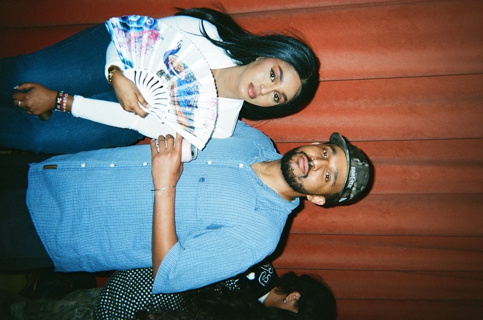 A young woman holding a decorative fan and standing next to a man wearing a hat, both posing for the photo in front of a wooden backdrop.
