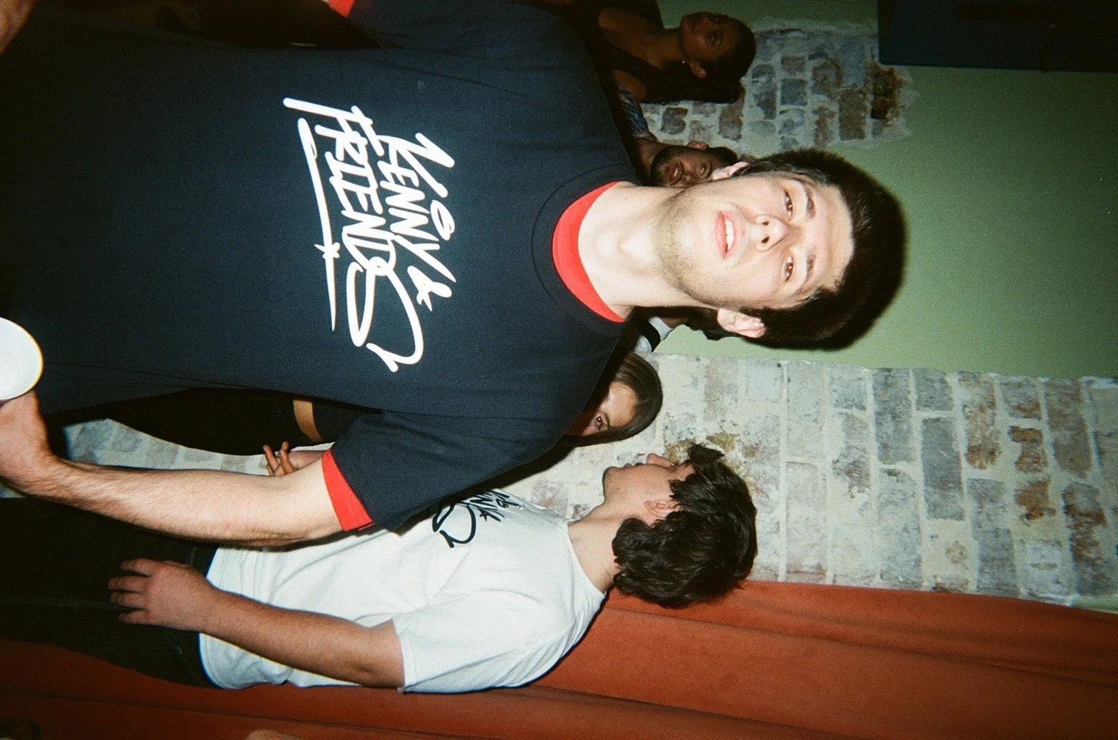 A group of young people at a social gathering, with one young man in the foreground wearing a black t-shirt with white text and a red collar, in front of a green wall with a stone section, and other young people in the background.