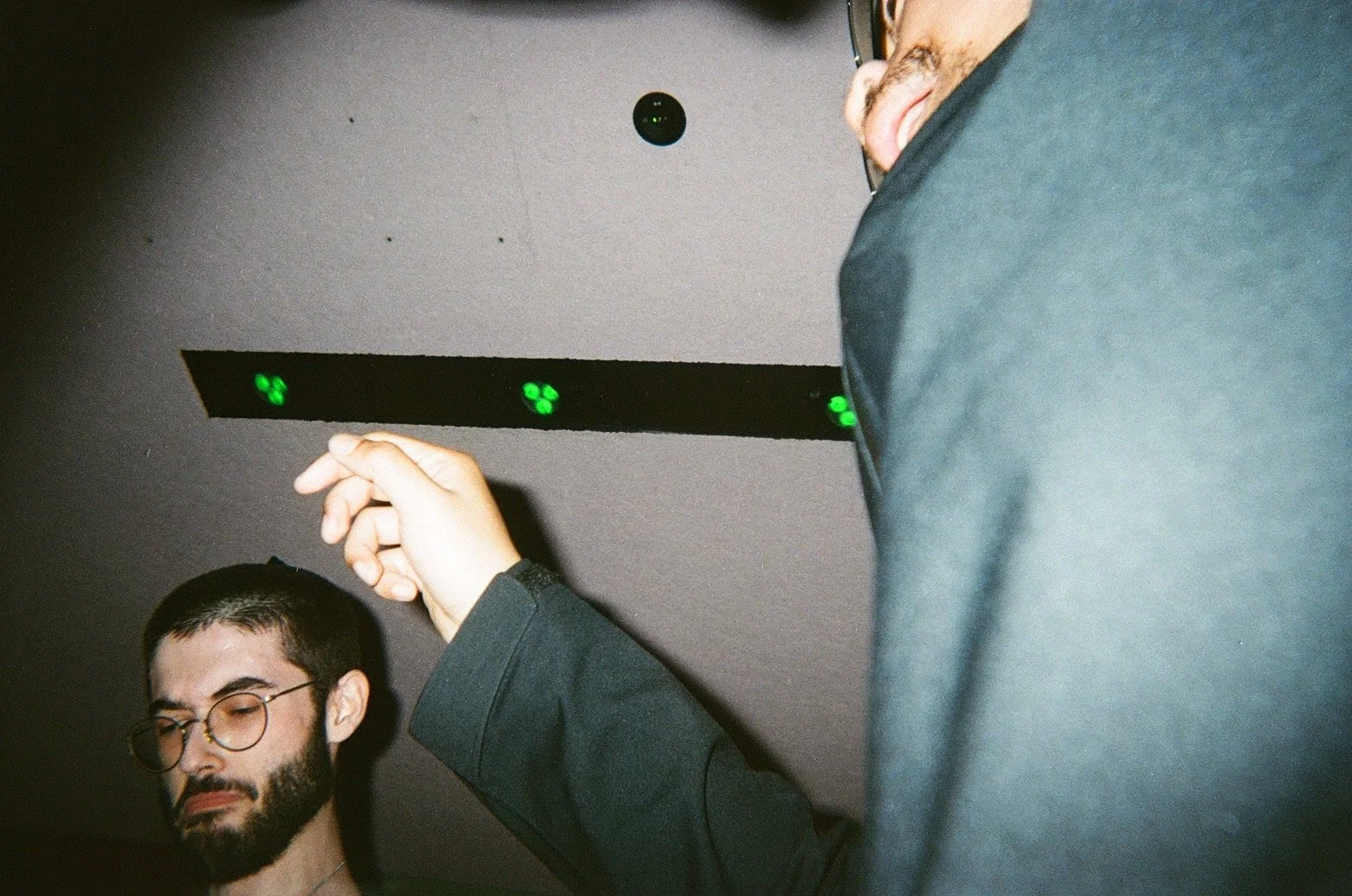Two men standing indoors near a ceiling with green lights and electronic equipment. One man with glasses and a beard, eyes closed, the other with glasses, partial face visible, appears to be adjusting something.