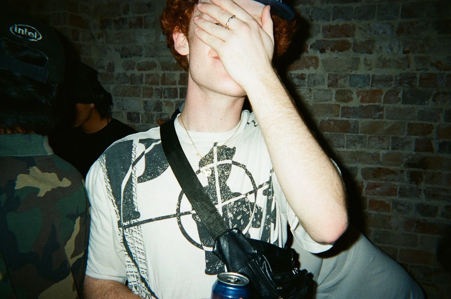 A young man with curly red hair covering his face with his hand, wearing a white graphic t-shirt, a gold necklace, and a black strap across his chest, holding a blue soda can, standing in front of a brick wall.