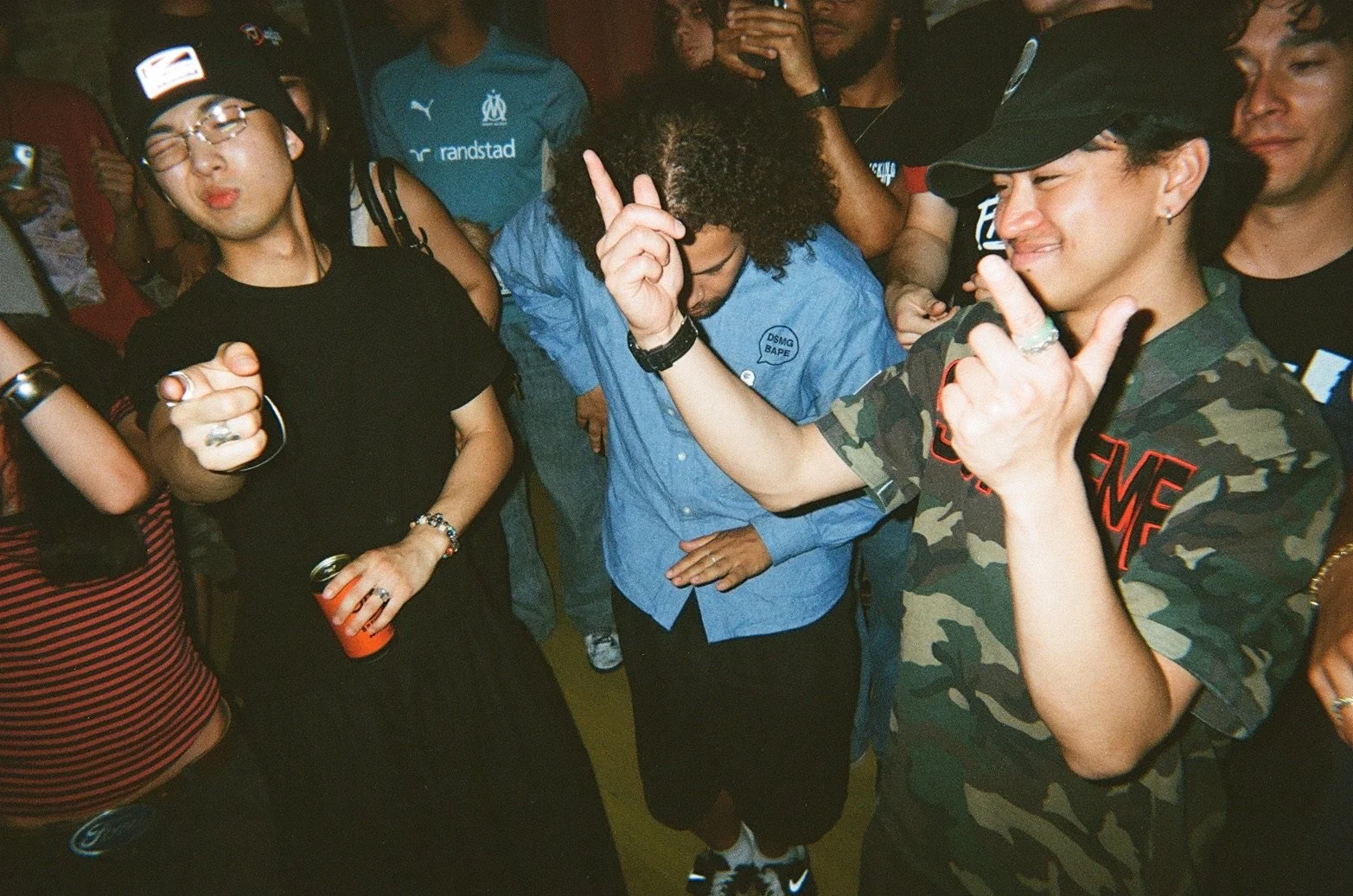 A group of people dancing and enjoying themselves at a party or social gathering. One person in the center with curly hair is making a gesture with their hand, while others around are smiling or holding drinks.