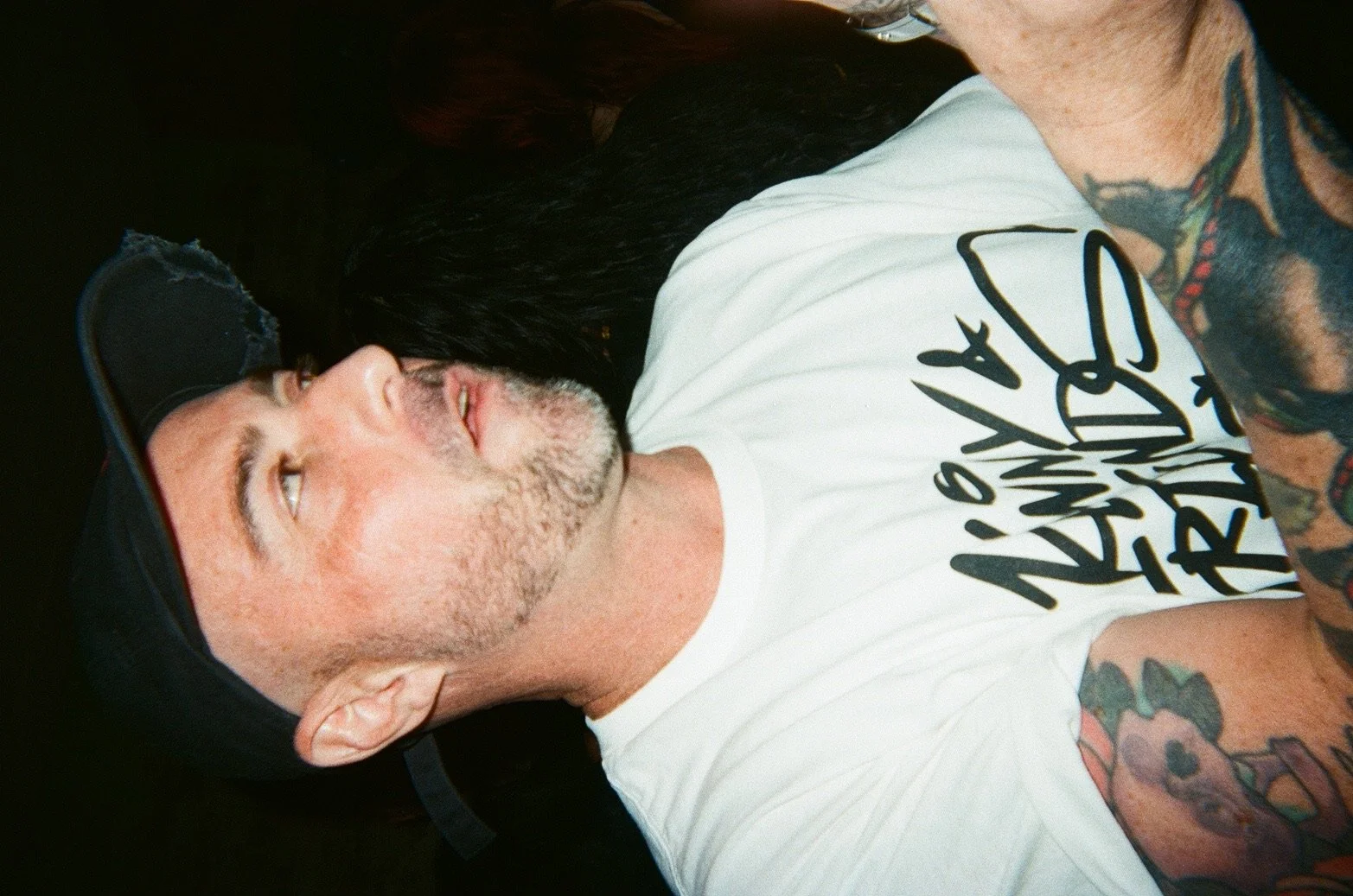 A man with tattoos wearing a black baseball cap, lying on his side with a white t-shirt, looking upwards in a dark setting.