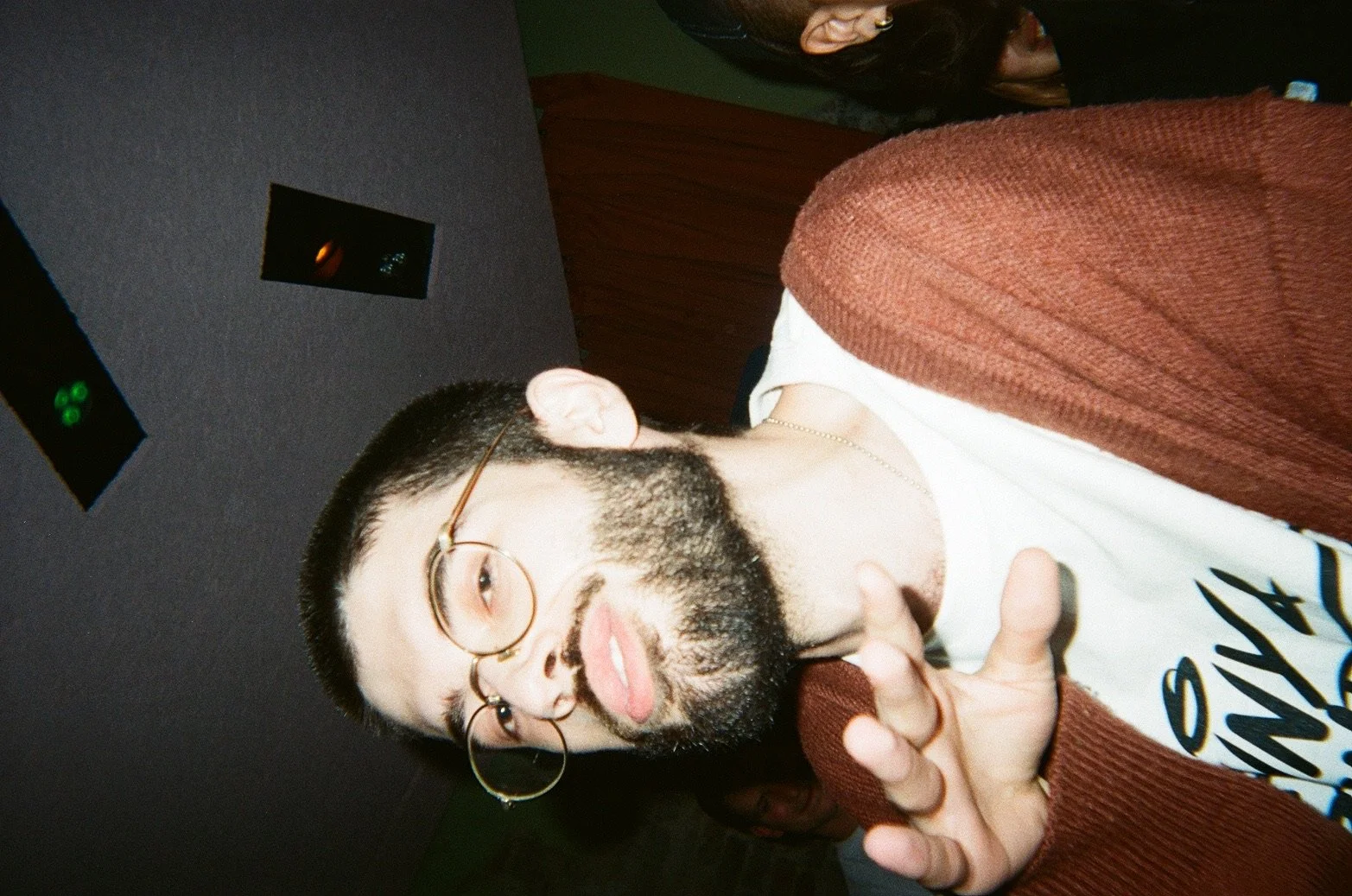 A man with glasses and a beard making a rock on hand gesture in a dimly lit indoor setting.