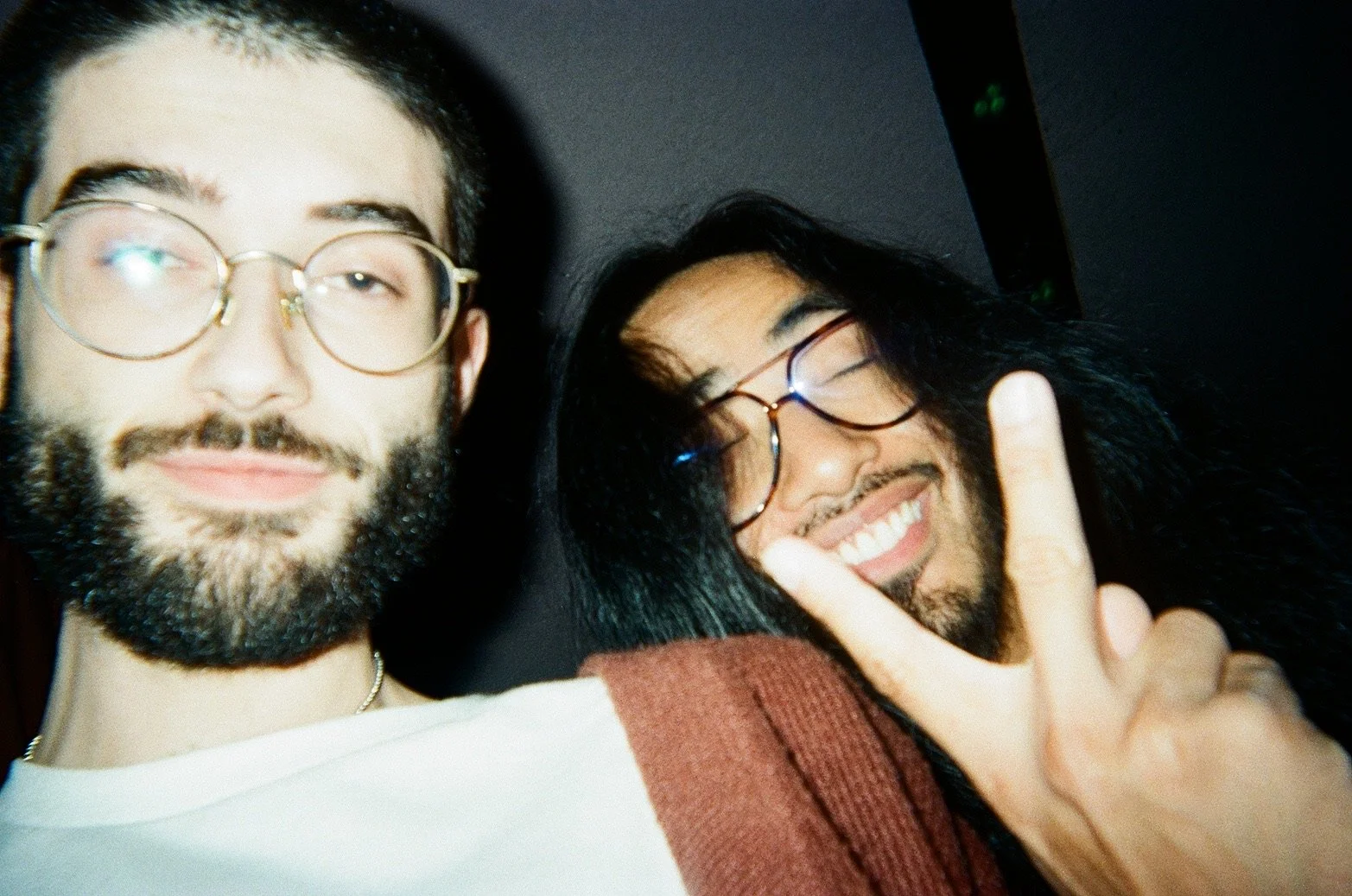 Two smiling men with long dark hair, beards, and glasses taking a close-up selfie. One man is making a peace sign with his hand.