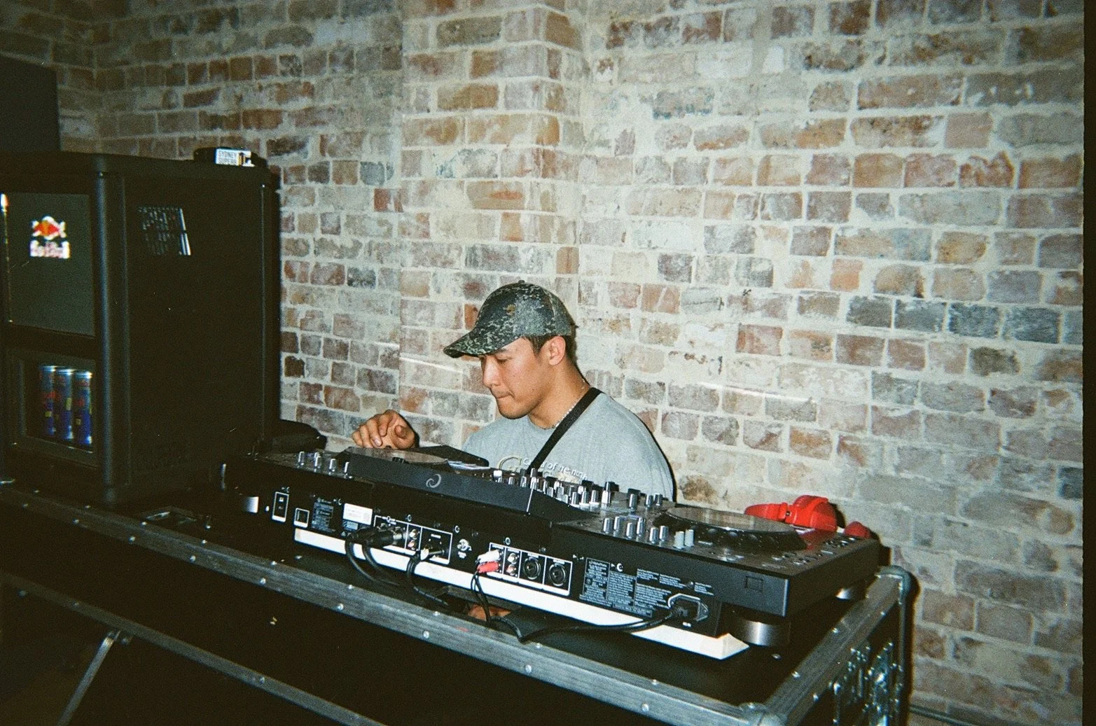 A DJ wearing a camouflage cap and a grey T-shirt stands behind a DJ mixer and turntable, concentrating on the equipment, against a rustic brick wall.