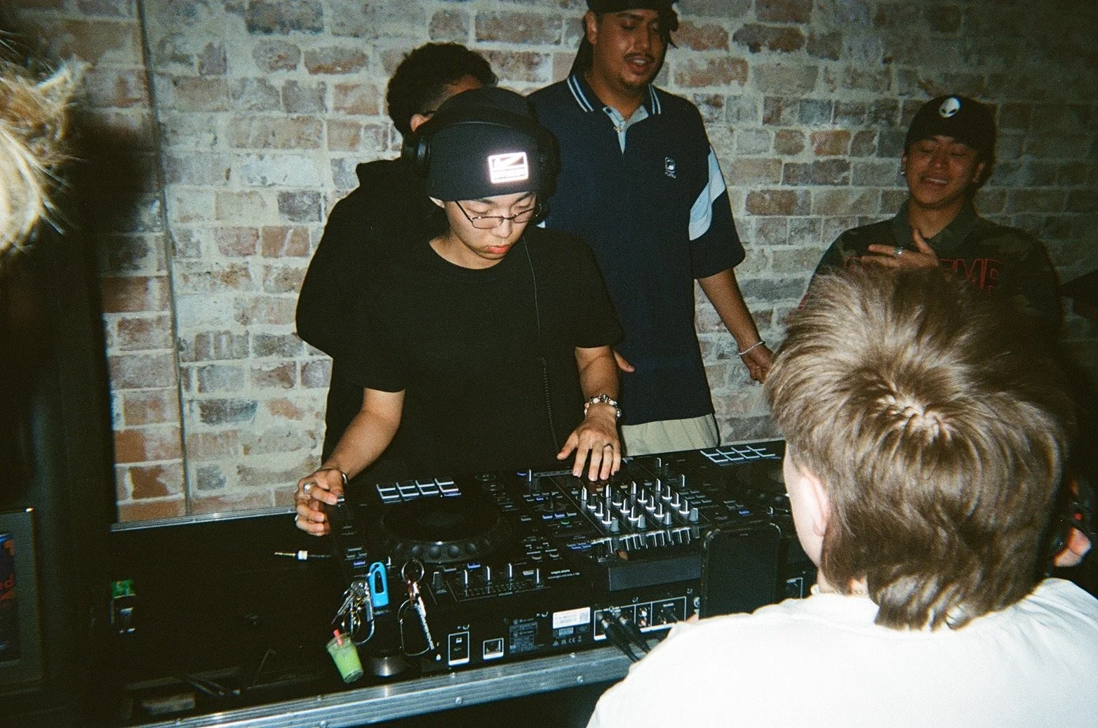 A woman DJ wearing headphones and glasses mixes music on a DJ controller in front of an audience, with a brick wall background.
