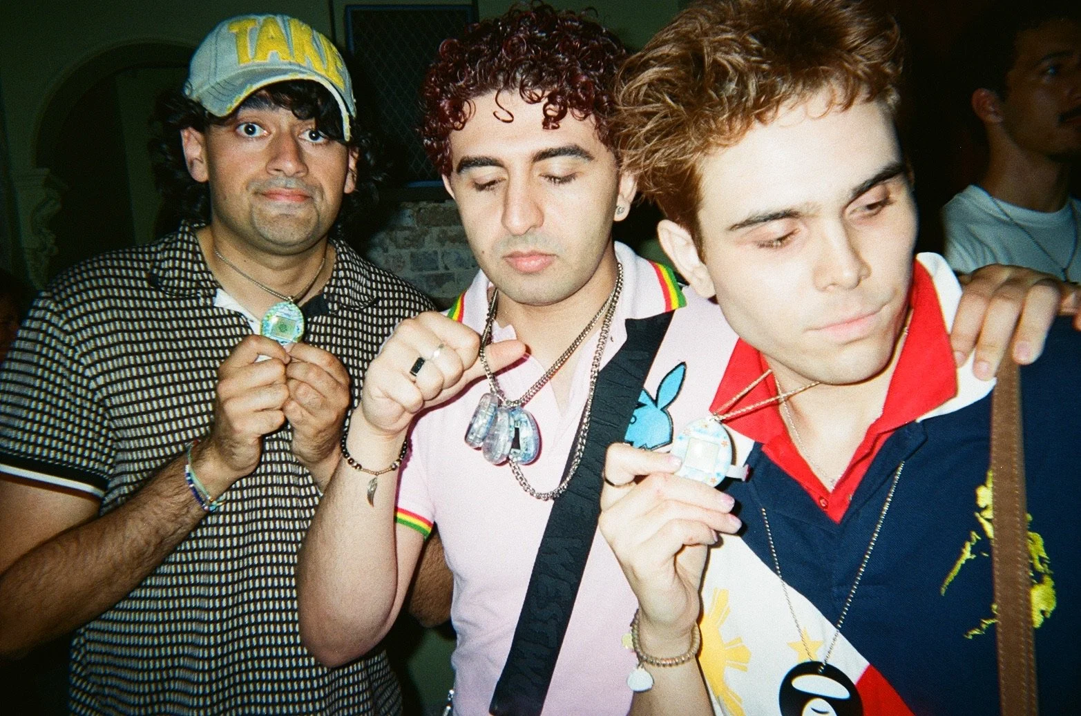 Three young men standing close together, showing off necklaces and pendants, with a fourth person partially visible in the background.
