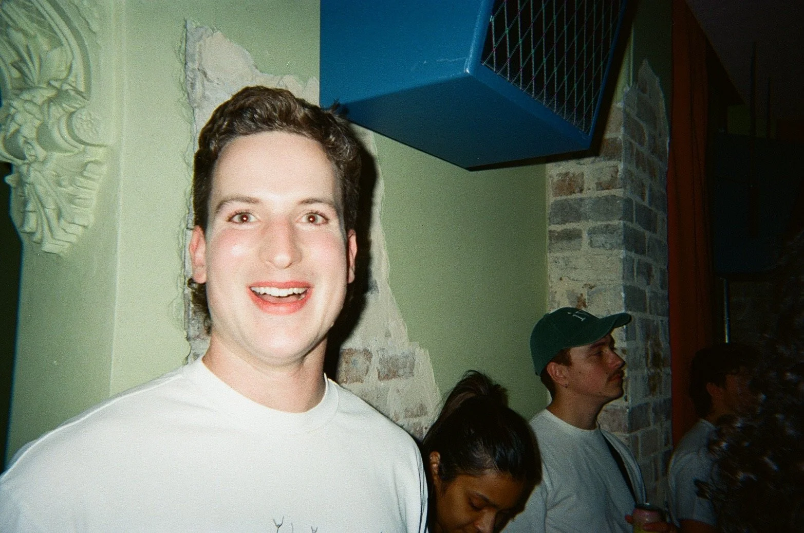 A smiling man with short dark hair wearing a white shirt, standing indoors against a wall with exposed bricks and a decorative relief, with other people in the background.