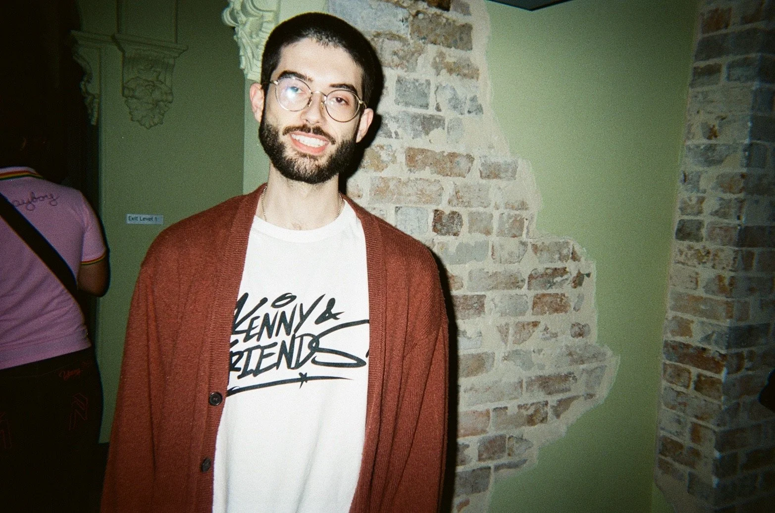 A young man with glasses, a beard, and dark hair smiling, wearing a white T-shirt with the words 'Kenny & Friends' and a red cardigan, standing in front of a brick wall and green painted wall.