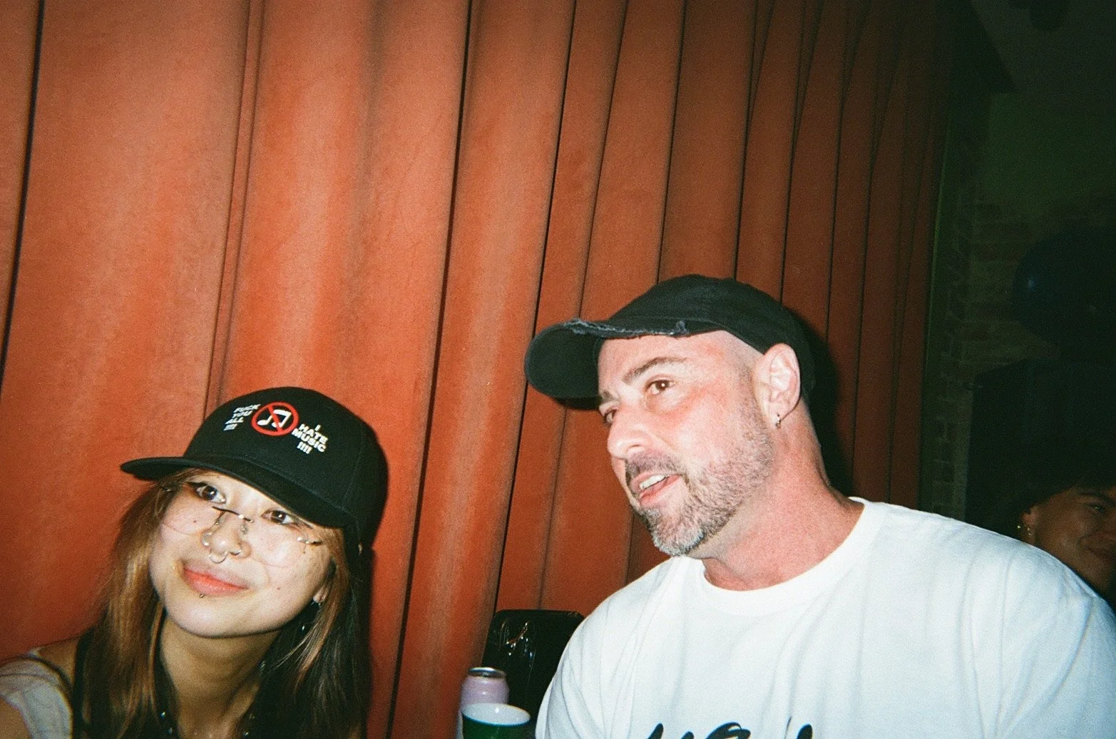 Two people, a woman with glasses and a man, are sitting together indoors. The woman is wearing a black cap with slogans and graphics, and the man is wearing a black cap and a white shirt. They are beside a red wooden wall, and two cans are visible on