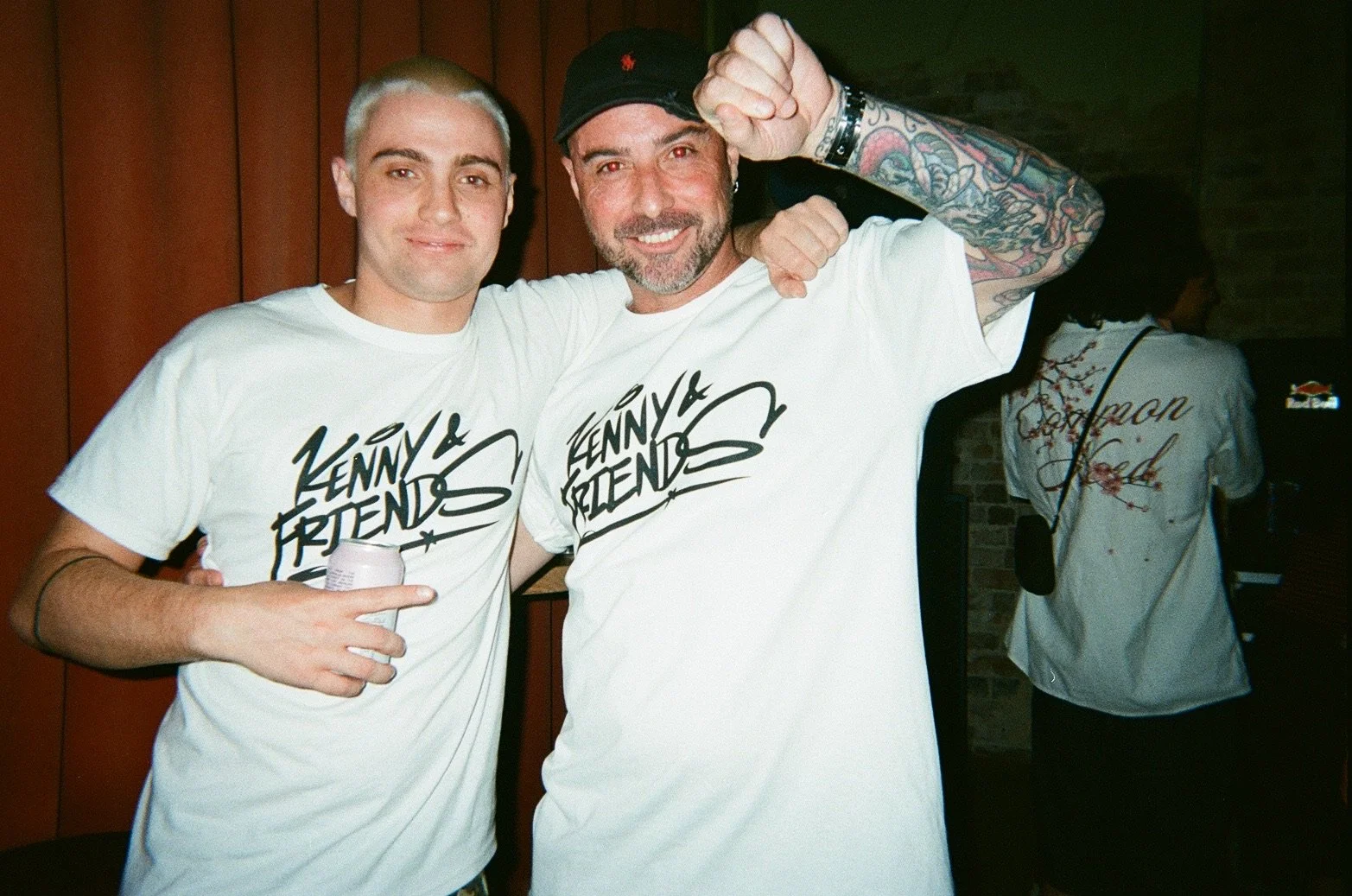 Two men wearing matching 'Kenny & Friends' T-shirts, smiling and posing for a photo indoors. One man has platinum blonde hair and is holding a drink, the other has dark hair, a beard, and colorful tattoos on his arm.
