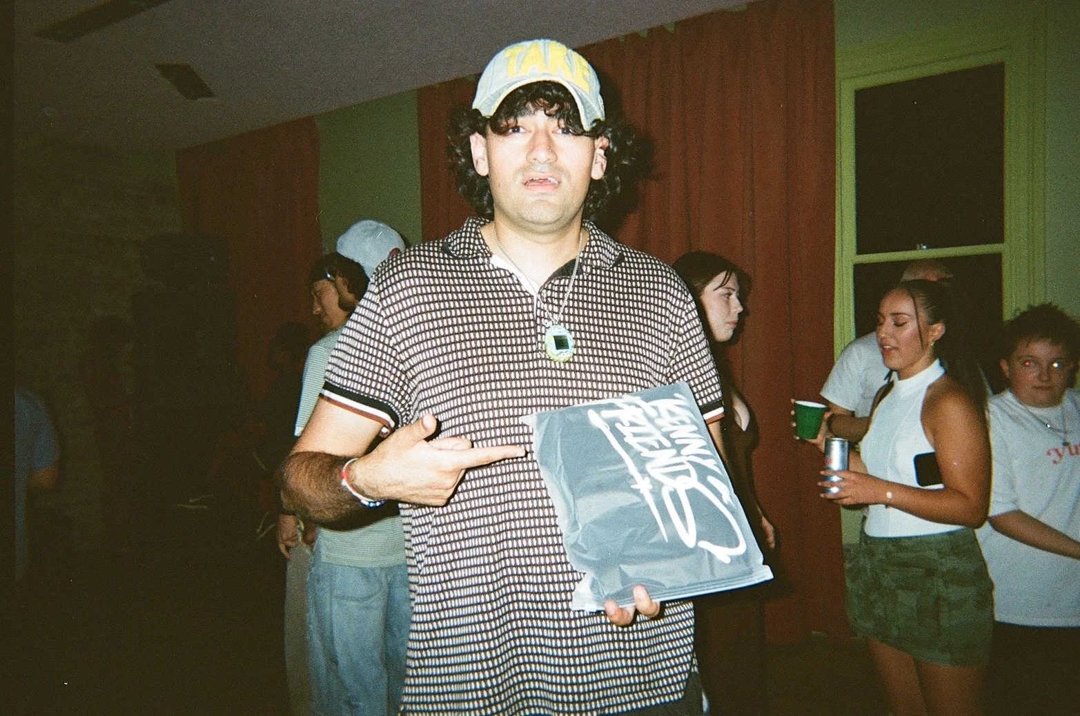 Young man with curly hair wearing a hat, pointing at his stomach, holding a plastic bag, at a party with others in the background.