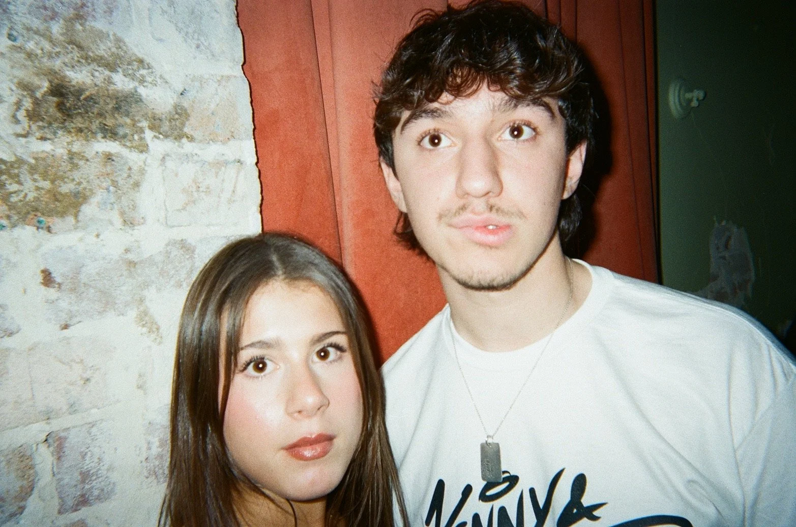 A young man and woman standing close together indoors. The woman has long brown hair and the man has dark wavy hair and a necklace. Behind them are a rustic brick wall and a wooden panel.