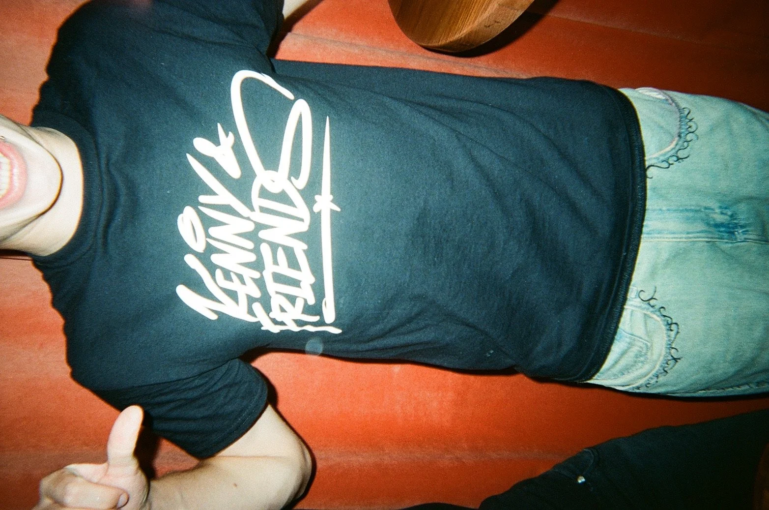 Person wearing a dark T-shirt with white graphic text, light-colored shorts, and giving a thumbs up, standing against a reddish wall.