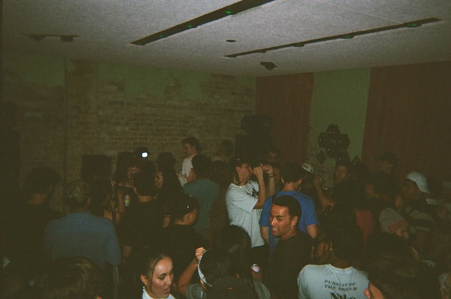 People at a crowded indoor party or event with exposed brick wall and a green wall, some are talking or interacting, one person is taking a photo with a phone, and others are smiling and enjoying themselves.