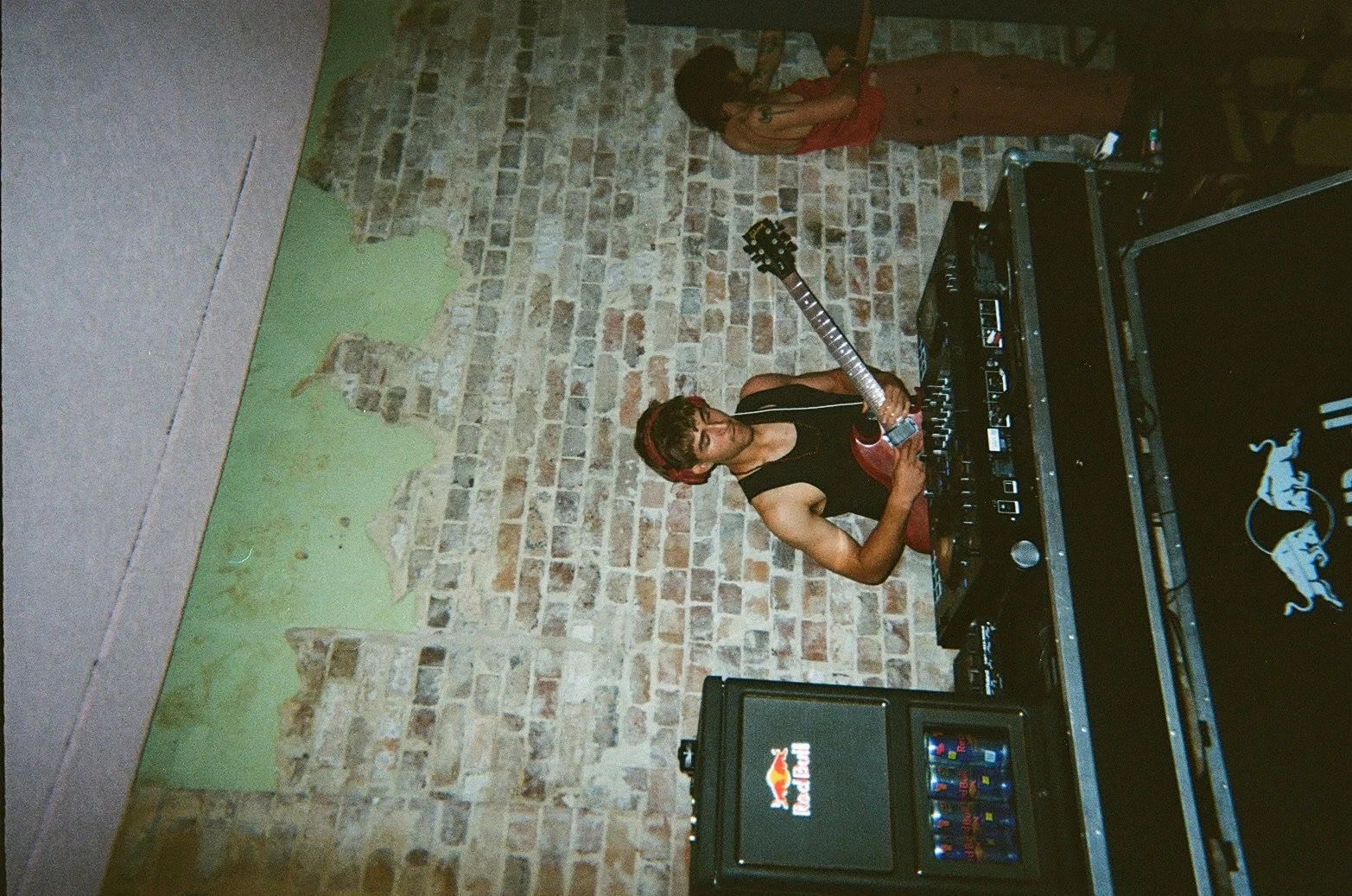 A woman with curly hair, a red headband, and black sleeveless top playing an electric guitar at a DJ setup in a room with brick and green walls.